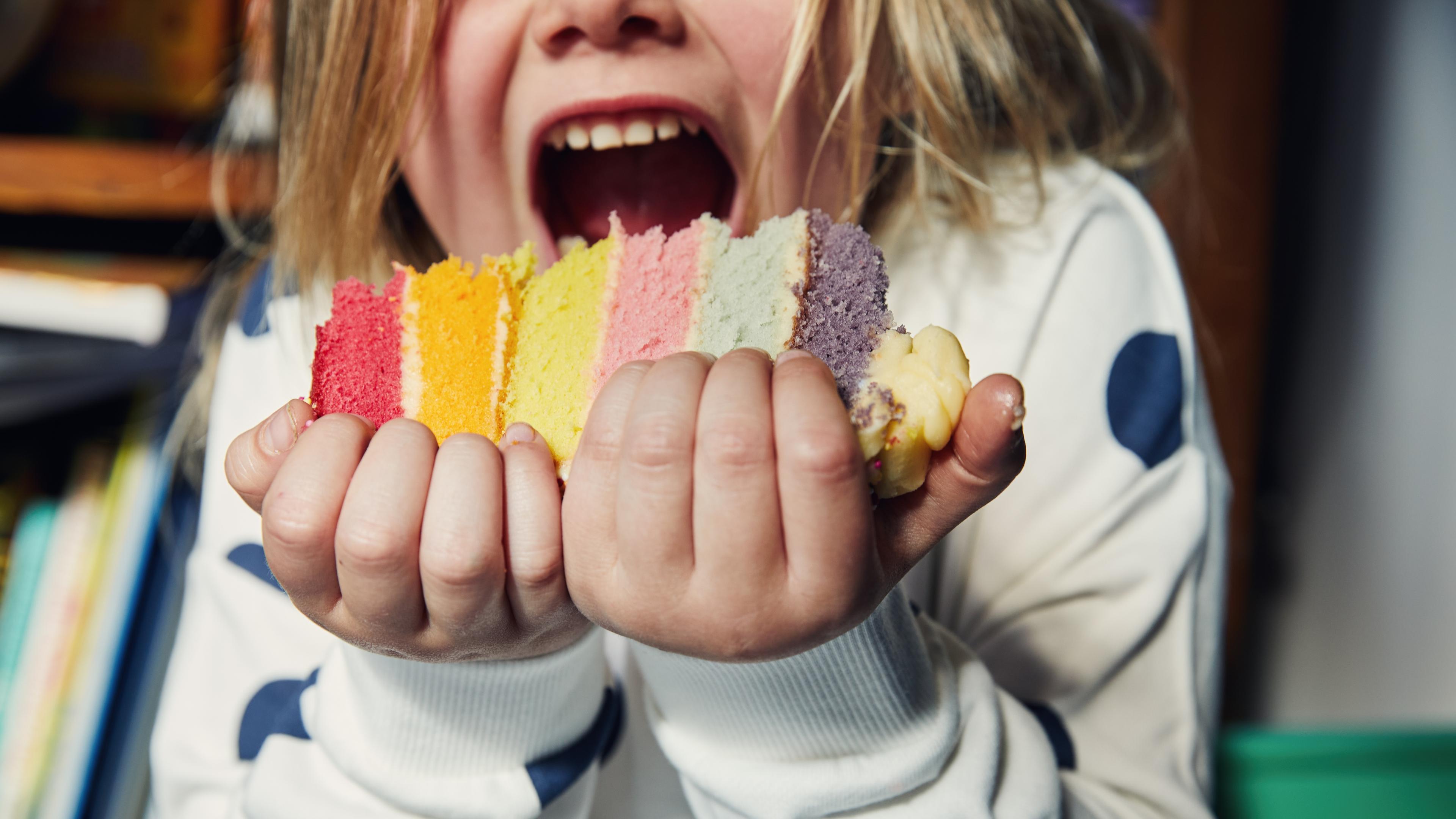 Photo of a child holding a colourful layered cake about to take a bite wearing a white jumper with blue spots.