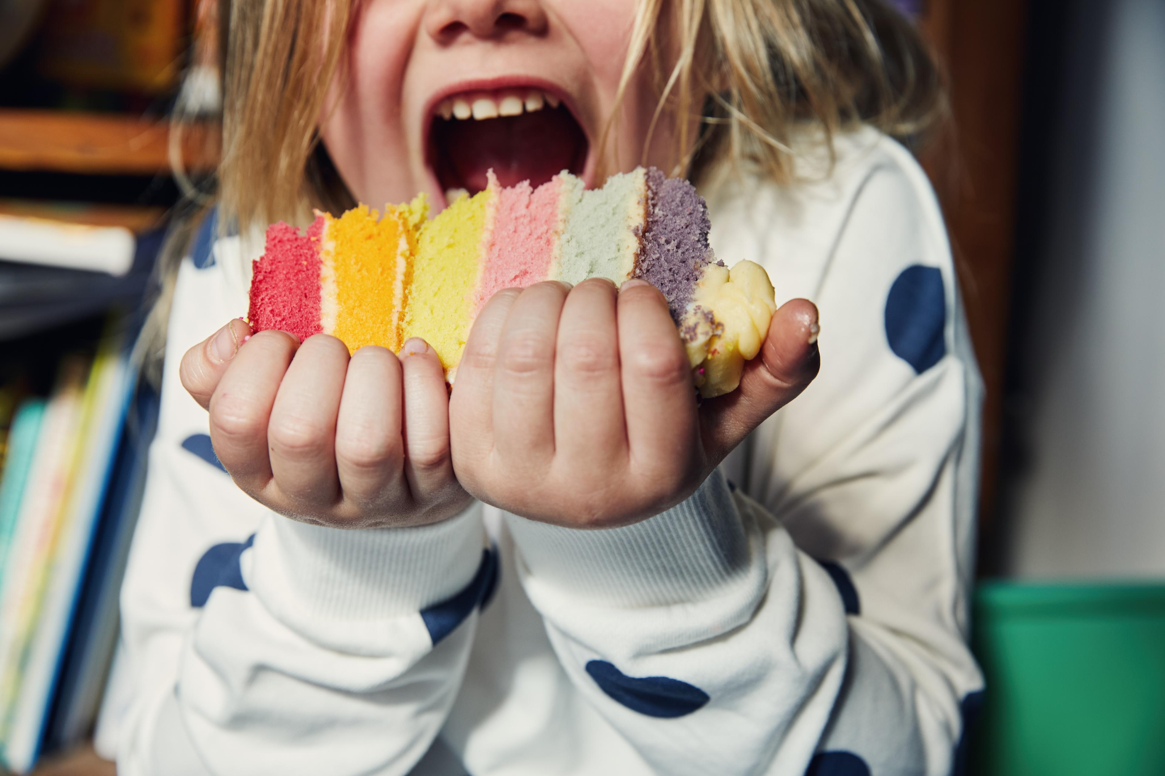 Photo of a child holding a colourful layered cake about to take a bite wearing a white jumper with blue spots.
