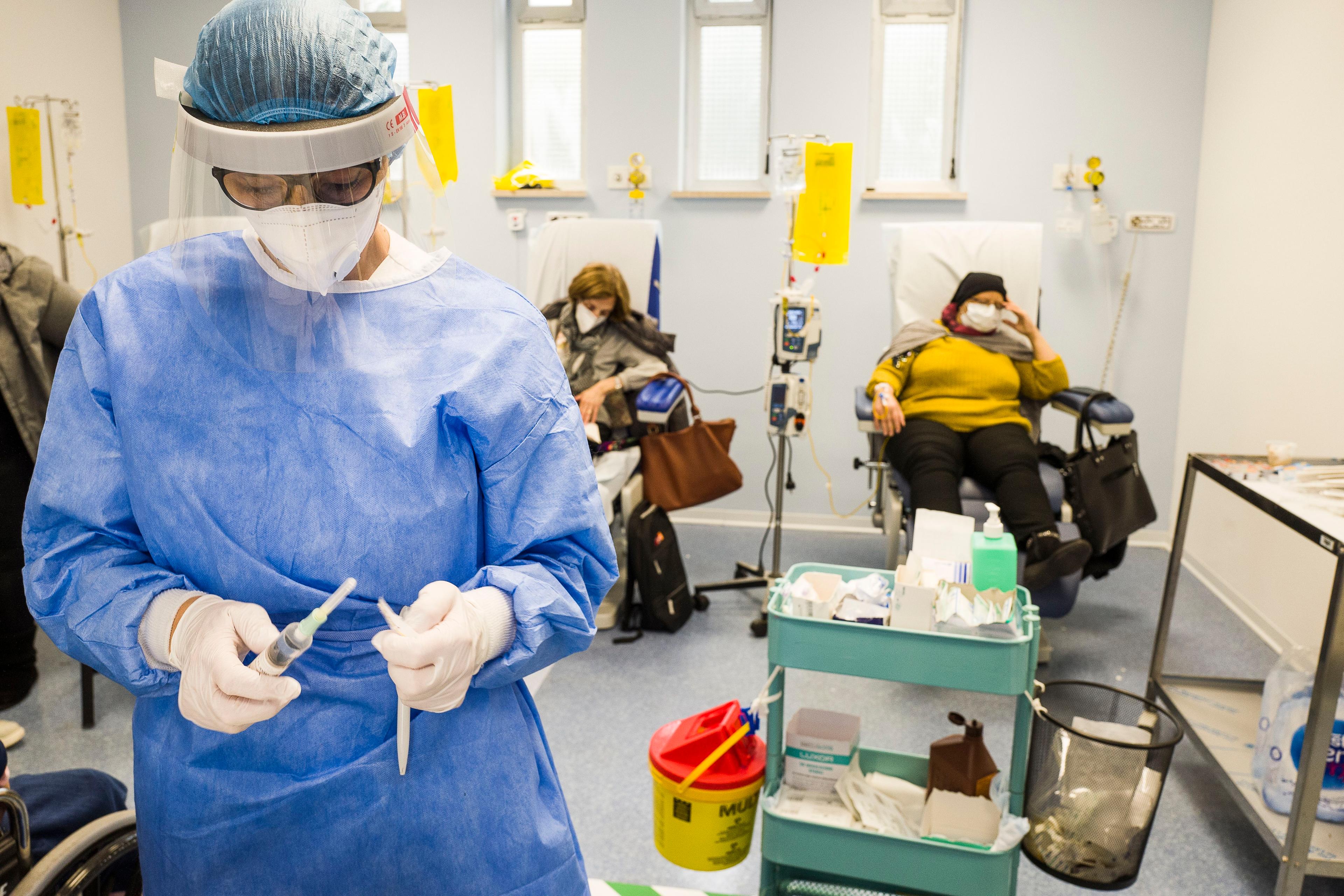 Photo of a healthcare worker in protective gear preparing a syringe in a hospital room with patients receiving treatment.
