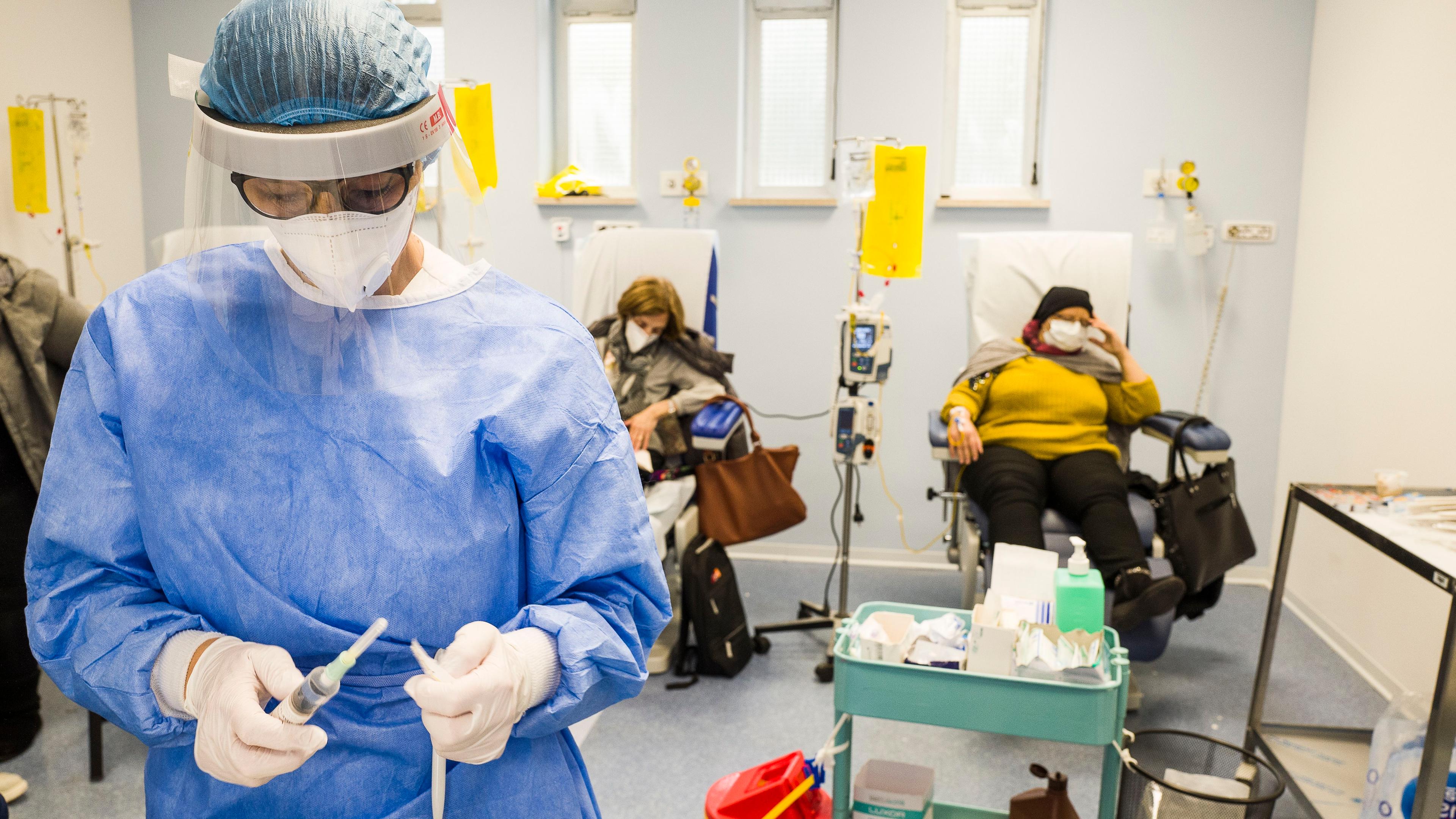 Photo of a healthcare worker in protective gear preparing a syringe in a hospital room with patients receiving treatment.