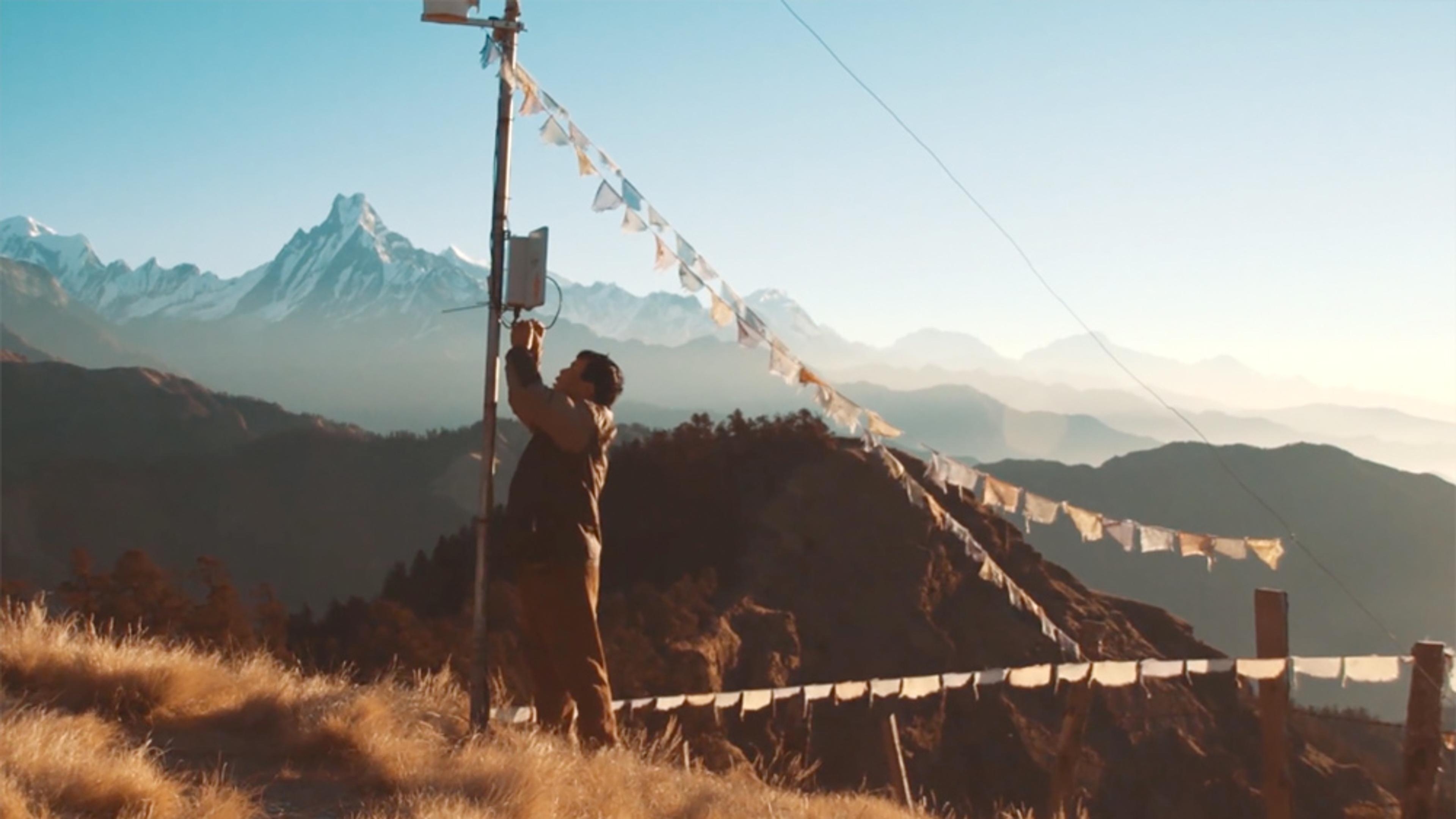 A person fixing equipment on a pole with prayer flags on a mountain, with snow-capped peaks and clear blue sky in the background.