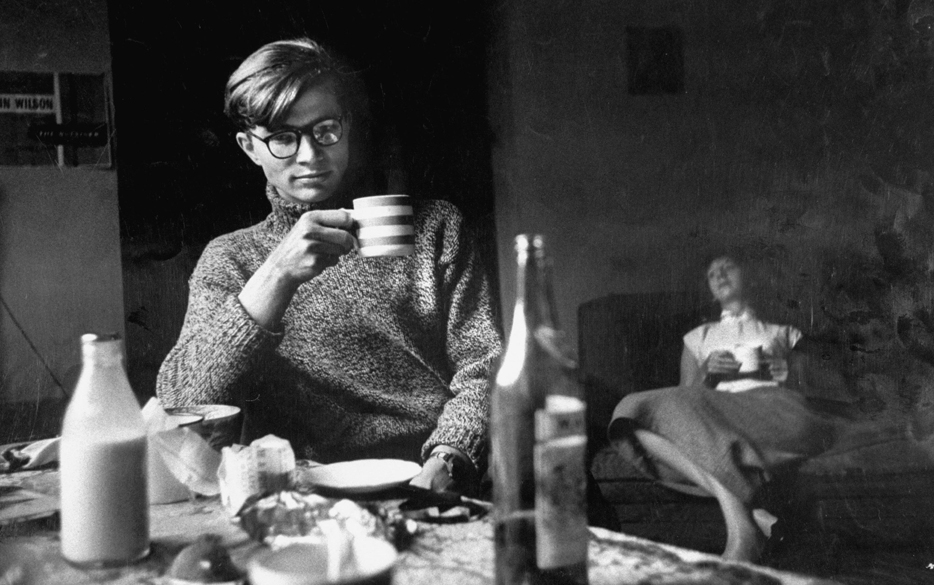 Black and white photo of a man in glasses drinking from a striped mug at a table with a woman seated in the background.