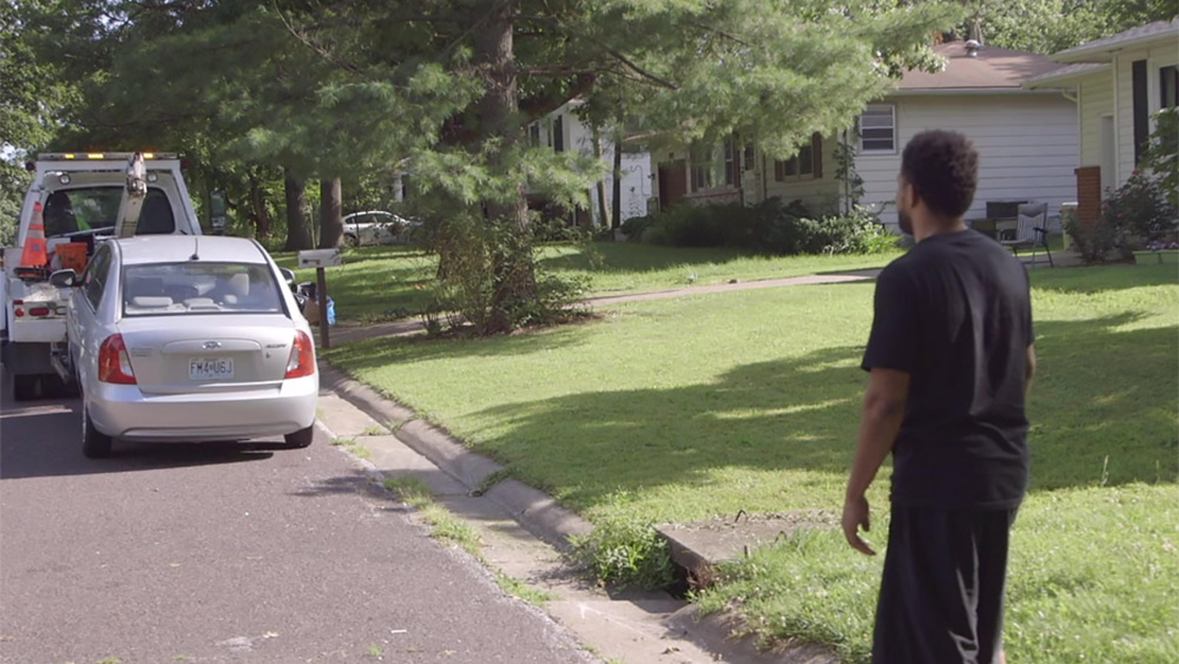 A man on a residential street watches a car being towed, with houses and a grassy lawn in the background.