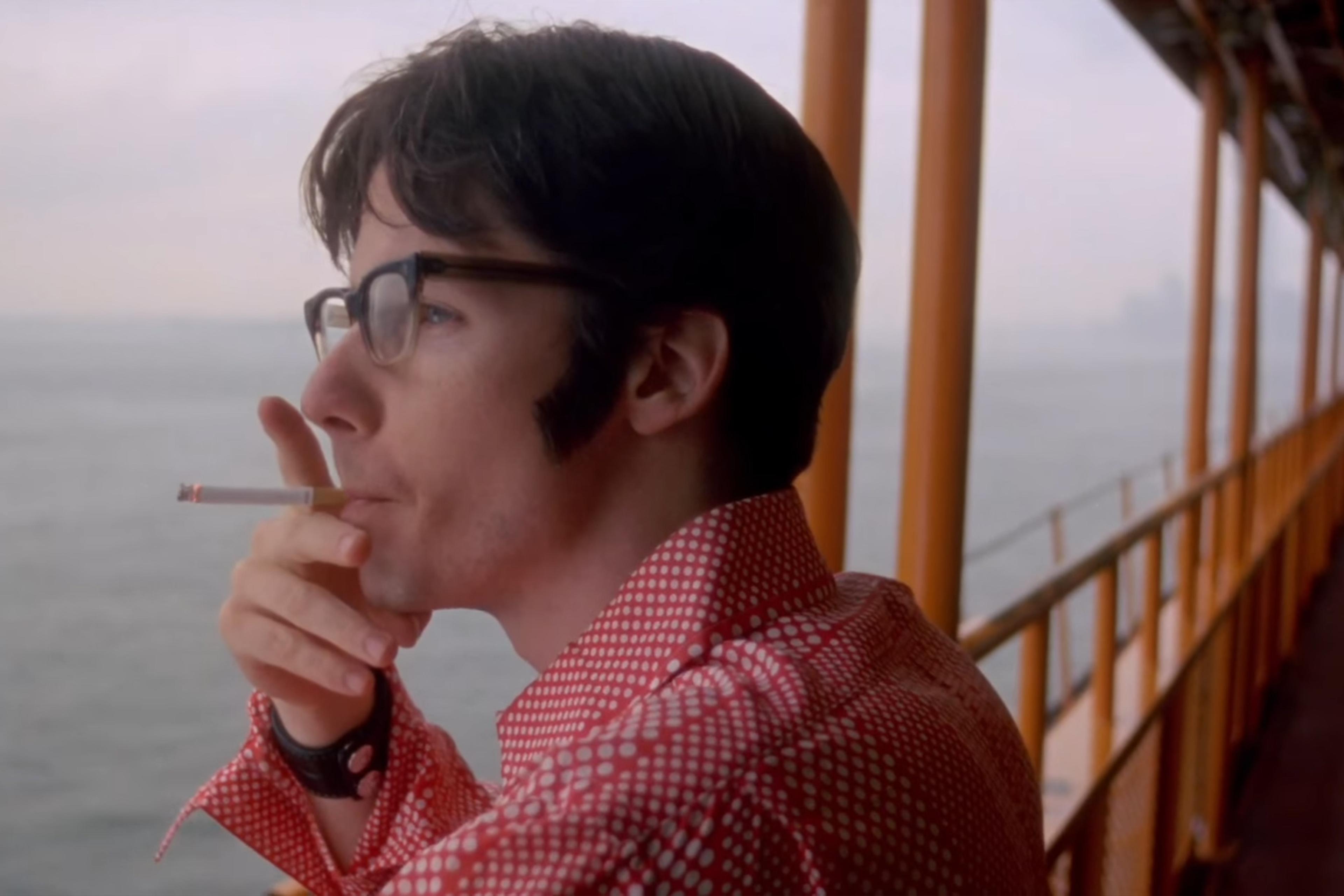 A man in glasses and a red polka dot shirt smokes a cigarette while gazing out over the water from a yellow railing.