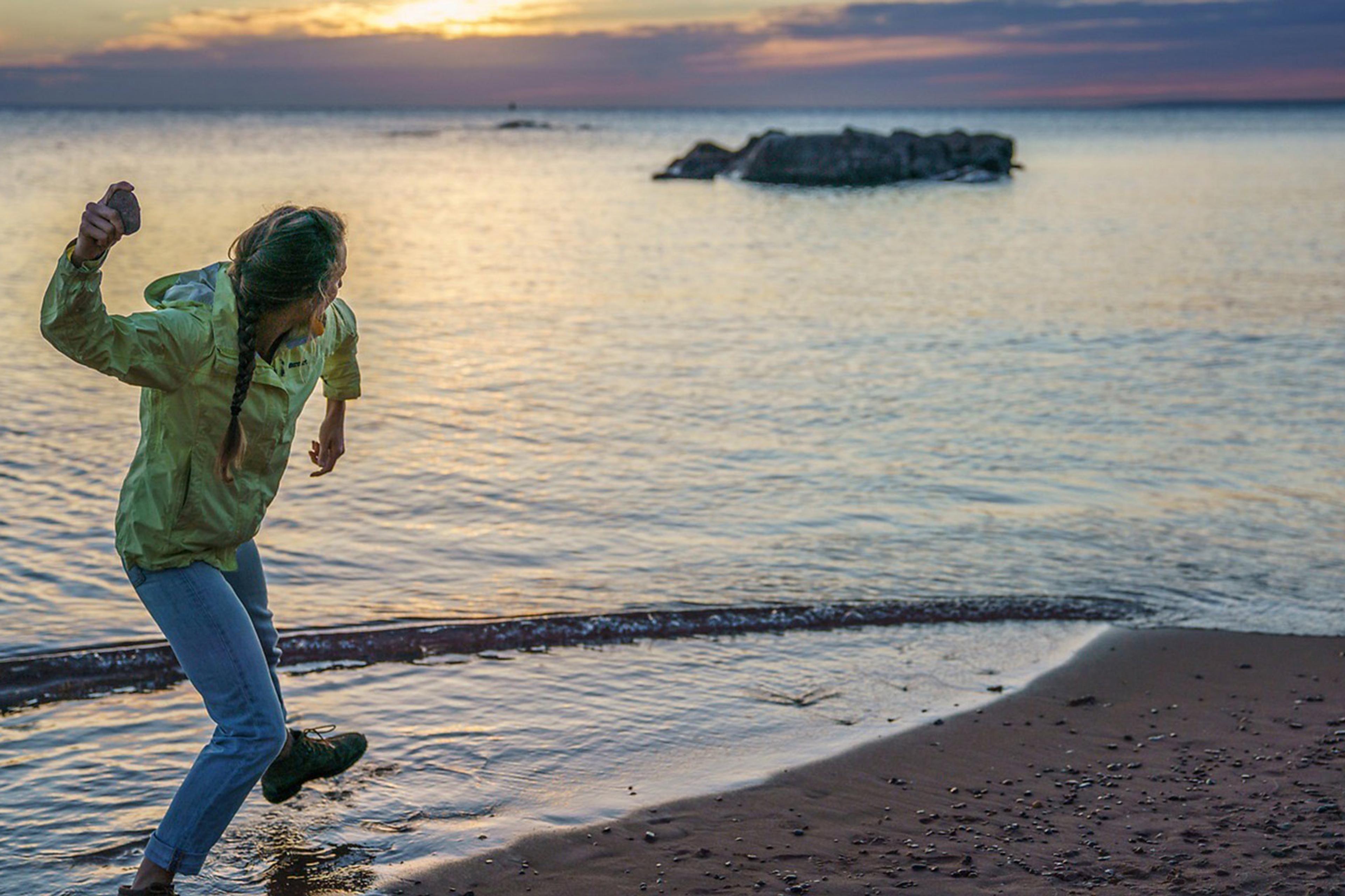 A person in a yellow jacket skipping stones at the beach during sunset, with calm water and rock formations in the distance.