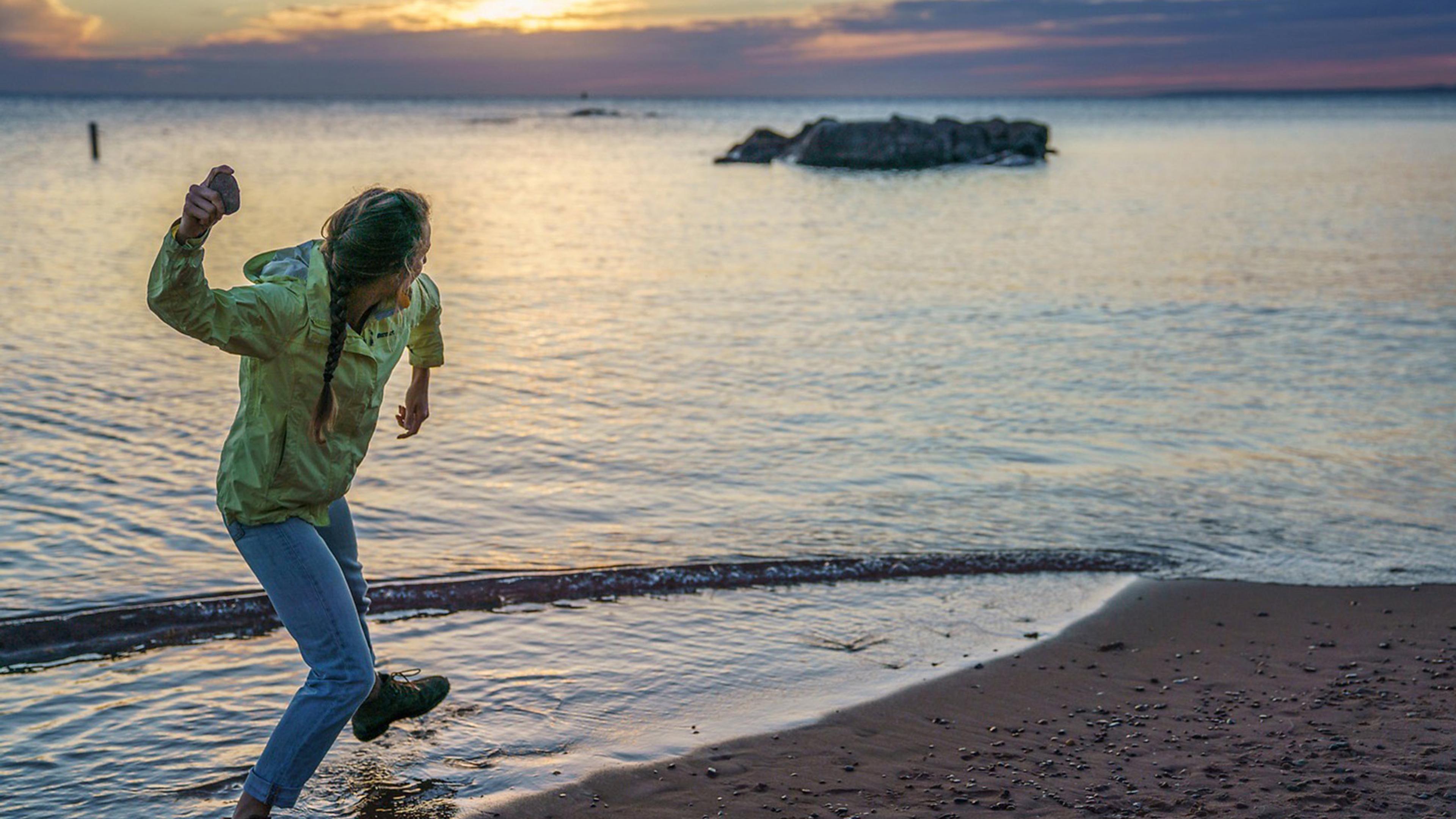 A person in a yellow jacket skipping stones at the beach during sunset, with calm water and rock formations in the distance.