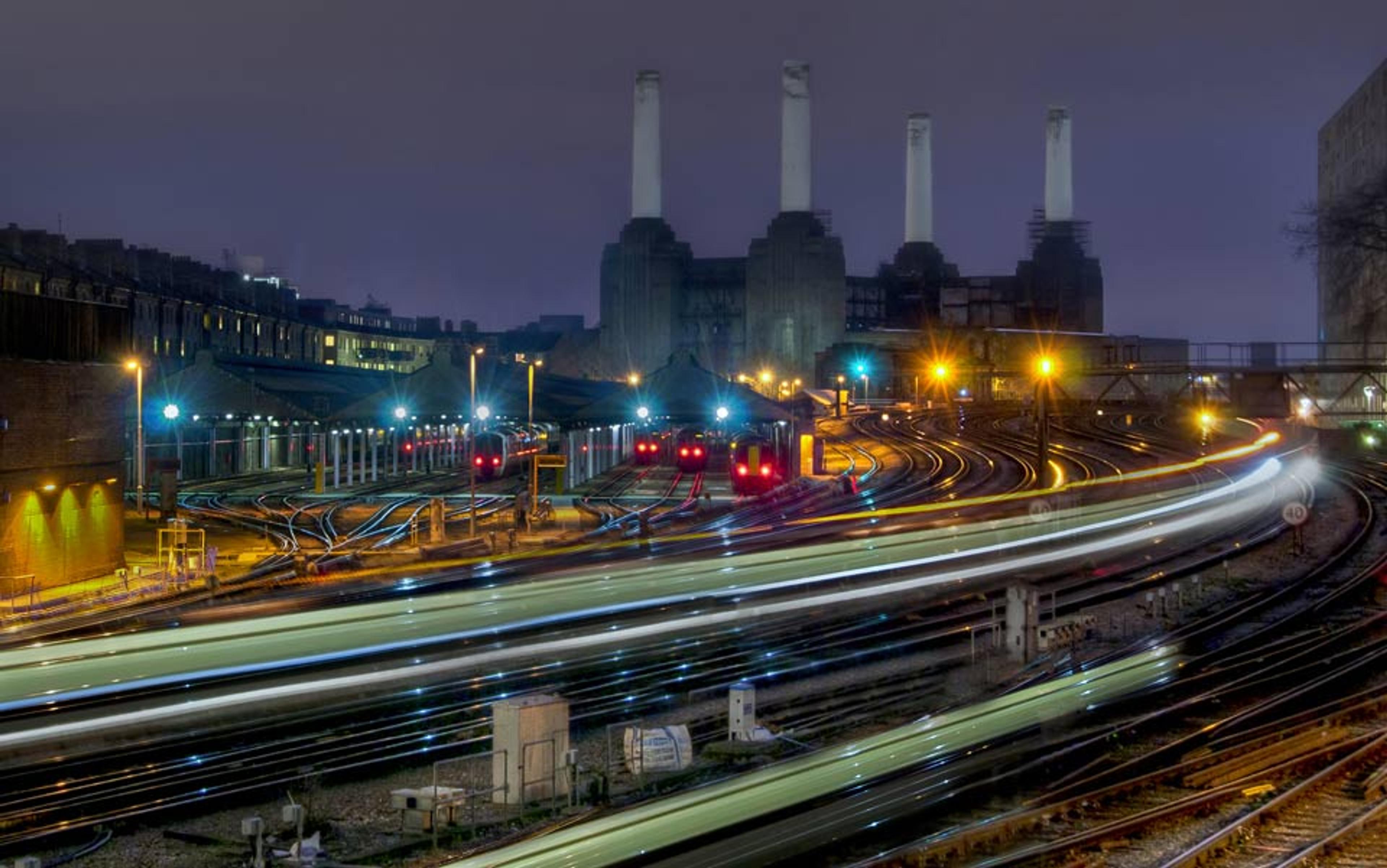 A railway yard at night with light trails from passing trains, illuminated signals and an industrial building in the background.