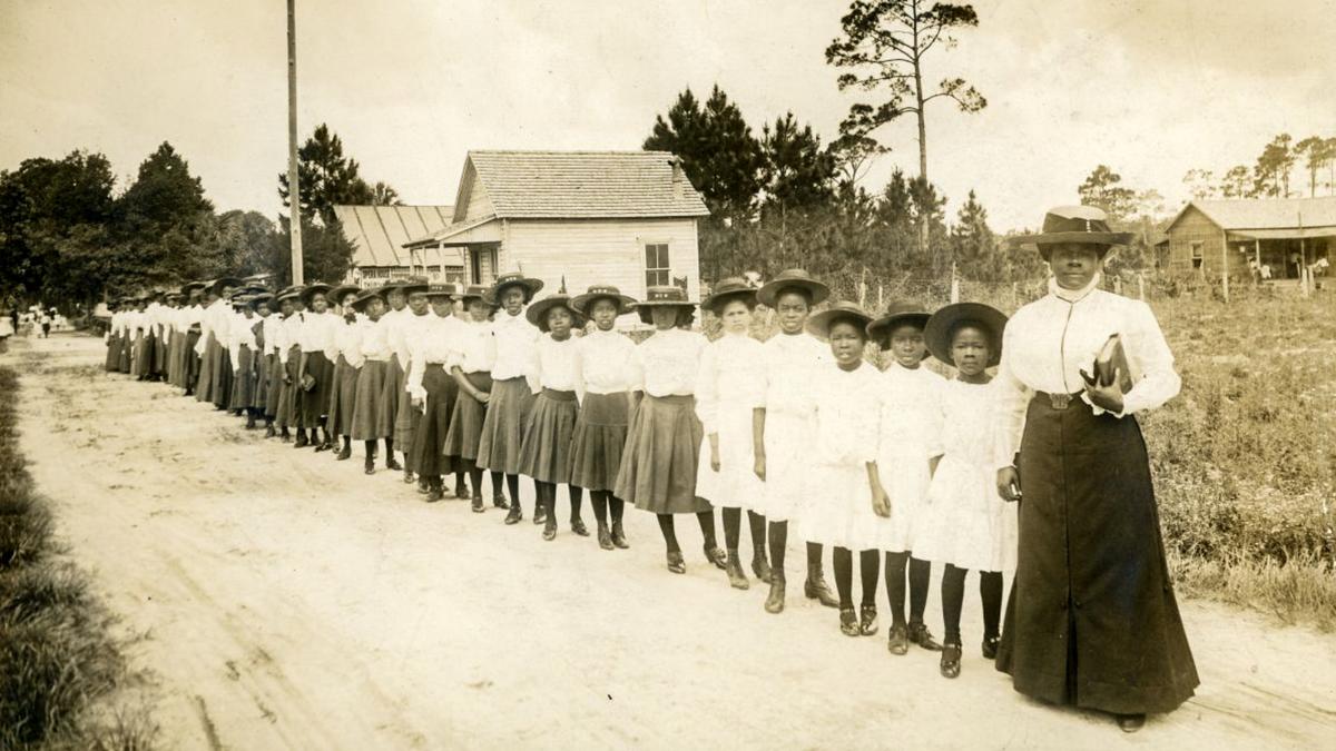 Historic photo of girls in hats and dresses standing in a line behind a woman on a dirt road beside wooden houses and trees.