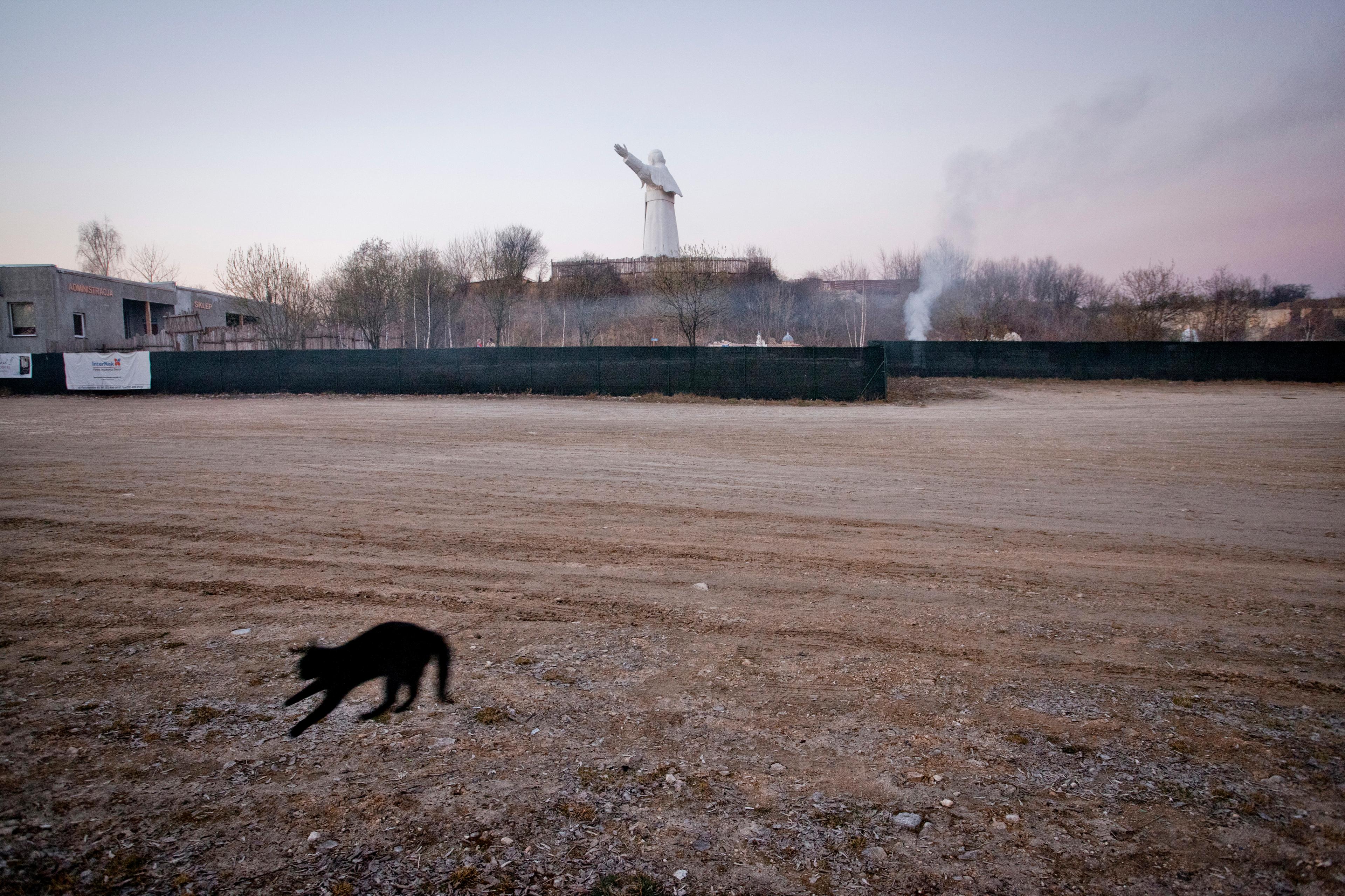 A blurred black cat jumps on dirt ground in the foreground, with a distant statue of Pope John Paul II with arms outstretched against the sky in the background