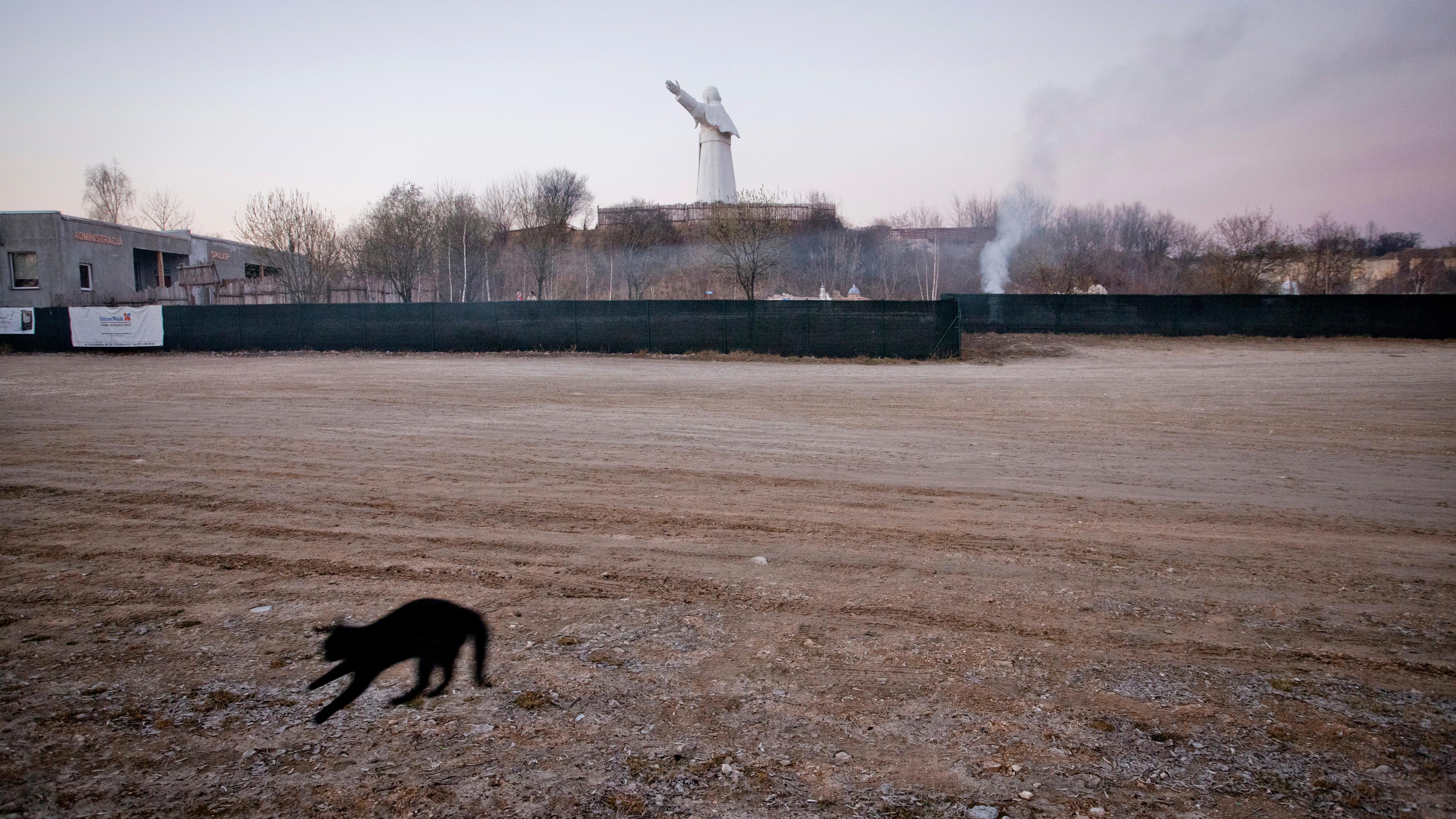 A blurred black cat jumps on dirt ground in the foreground, with a distant statue of Pope John Paul II with arms outstretched against the sky in the background