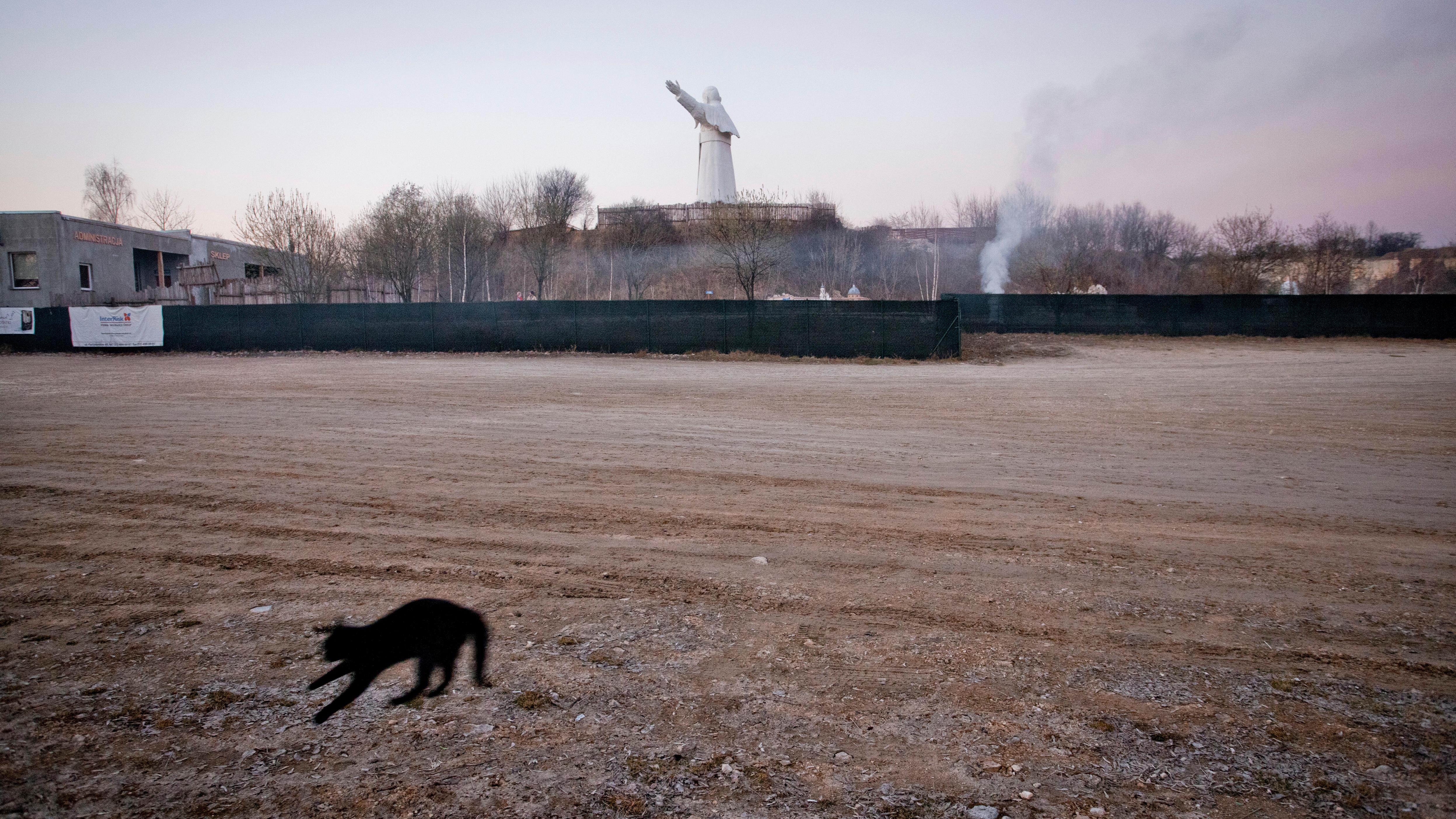 A blurred black cat jumps on dirt ground in the foreground, with a distant statue of Pope John Paul II with arms outstretched against the sky in the background