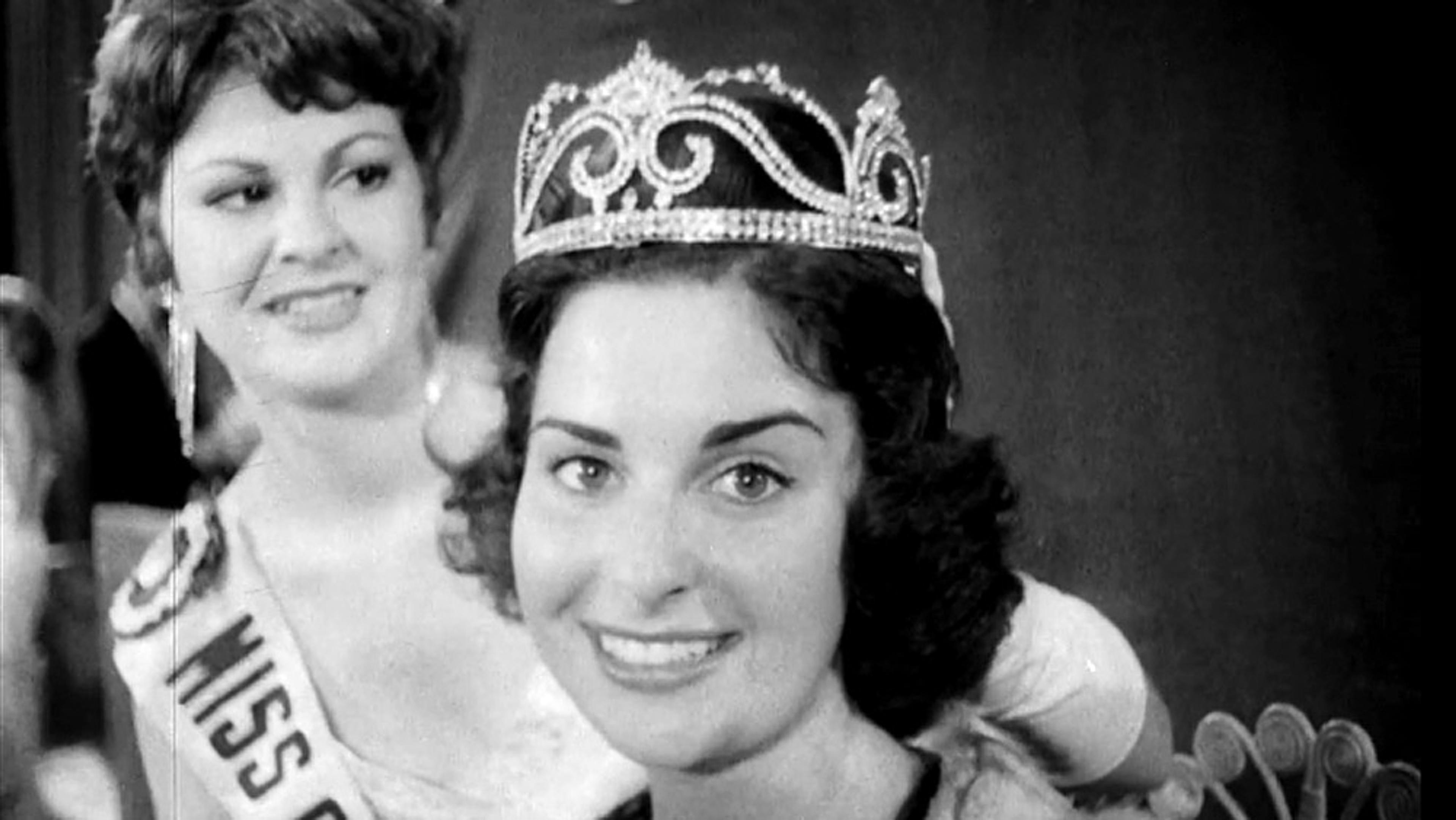 Black and white photo of a smiling woman wearing a crown next to another woman with a sash reading “Miss”.