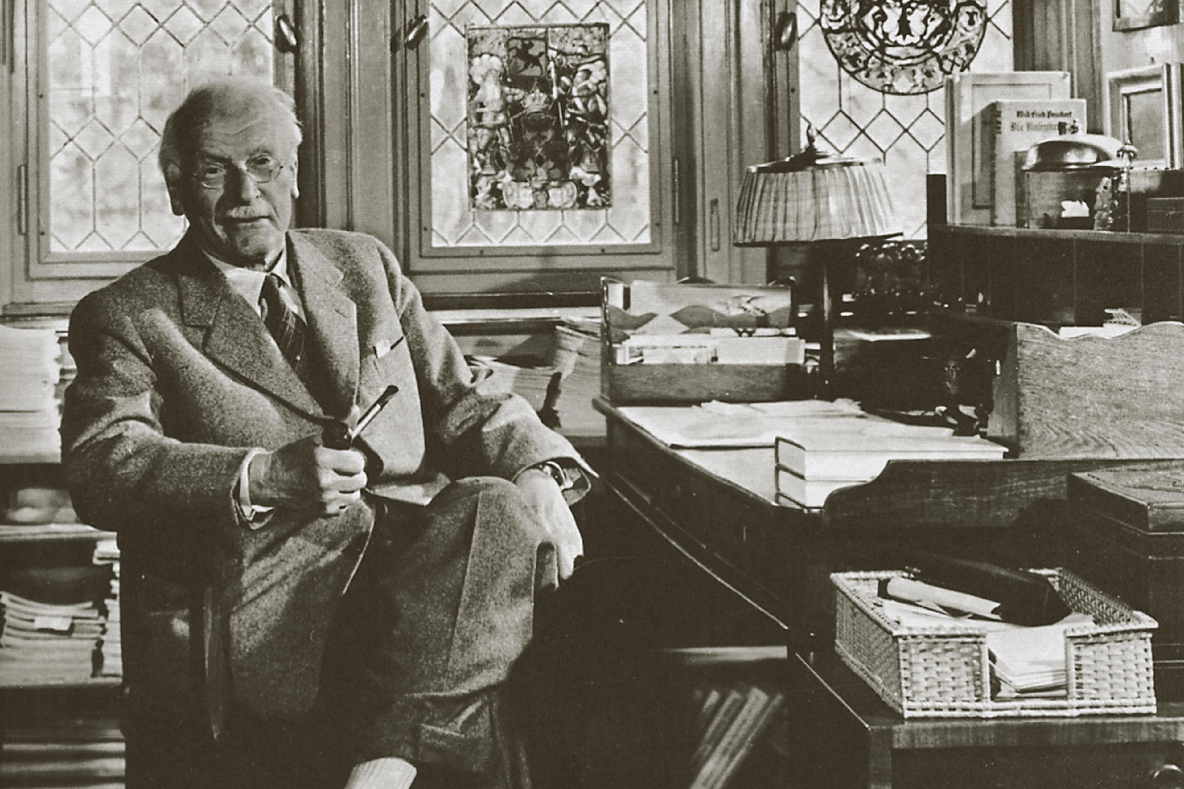 Black-and-white photo of a man with glasses holding a pipe, sitting in an office with stained glass windows.