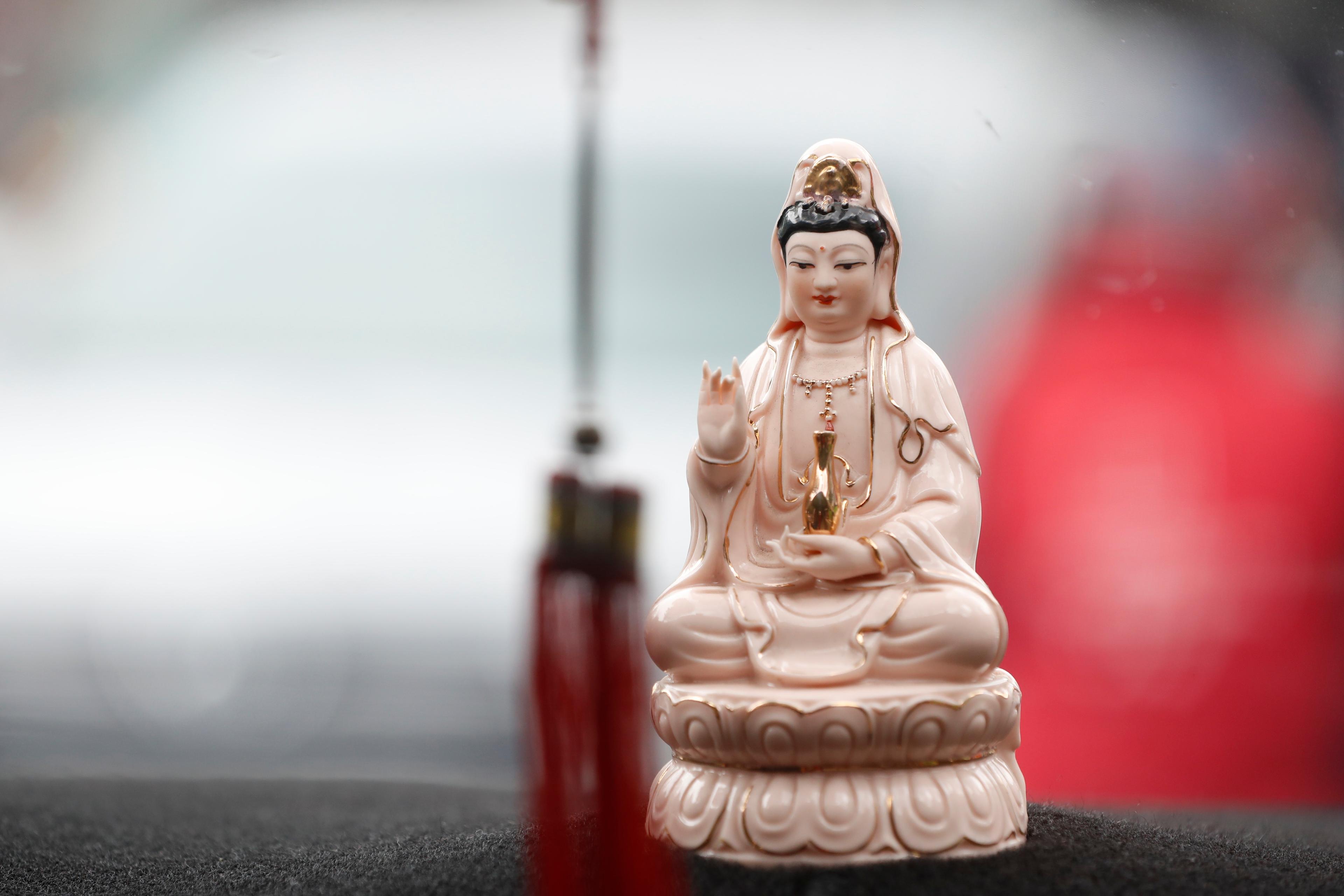 Photo of a small porcelain statue of a serene figure in robes, holding a vase on a dashboard with a blurred background.