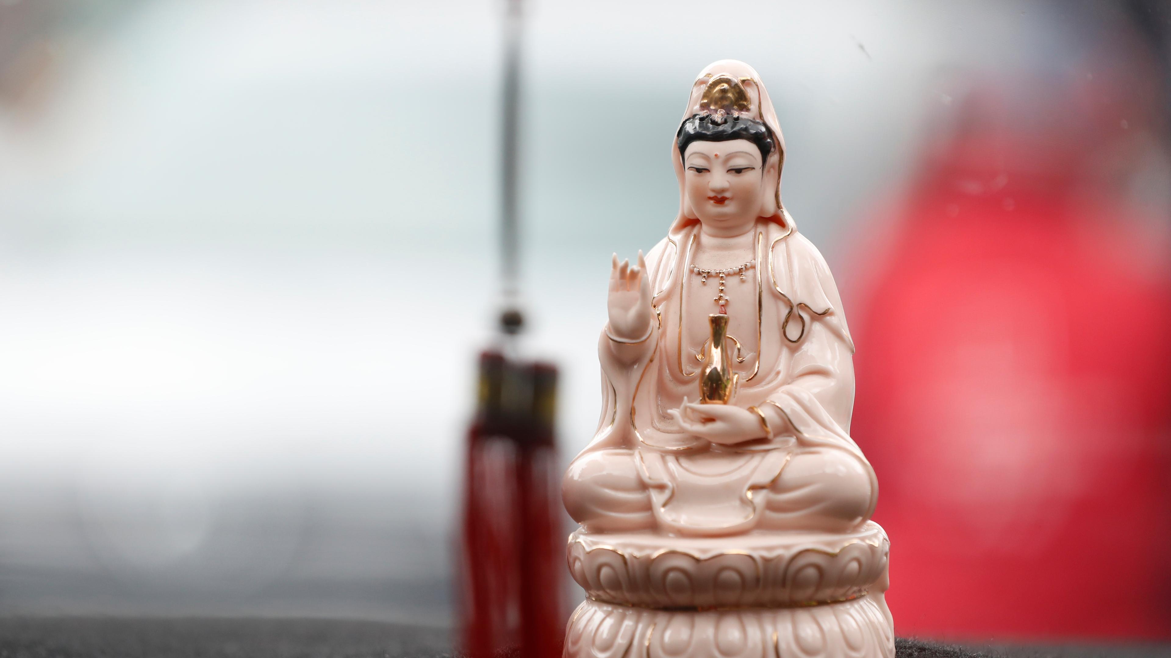 Photo of a small porcelain statue of a serene figure in robes, holding a vase on a dashboard with a blurred background.