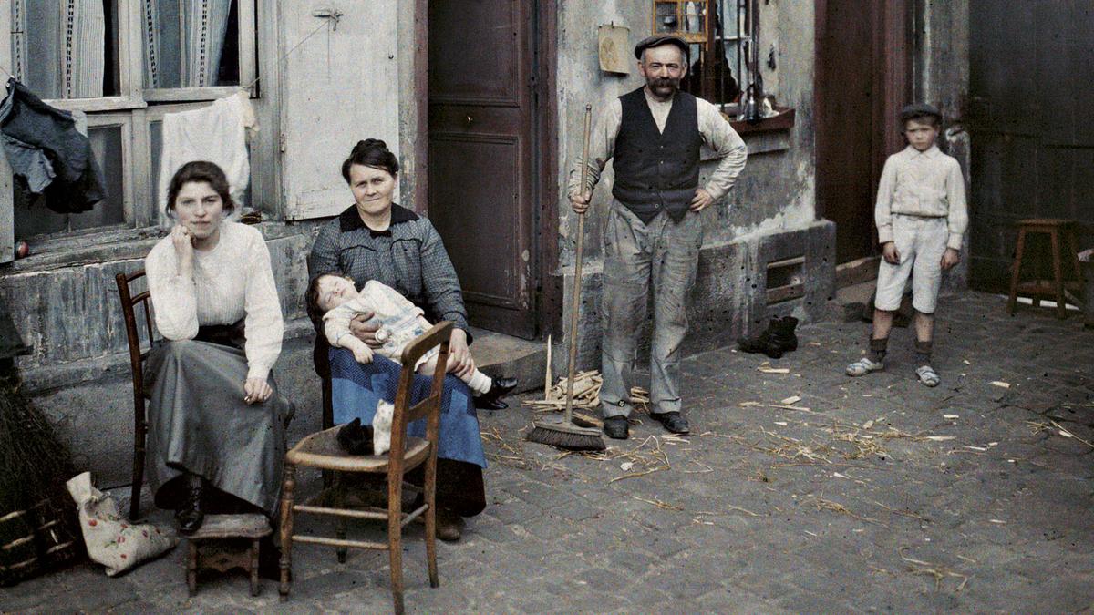 Early 20th-century colour photo of a family sitting and standing outside a rustic building on a cobblestone street.