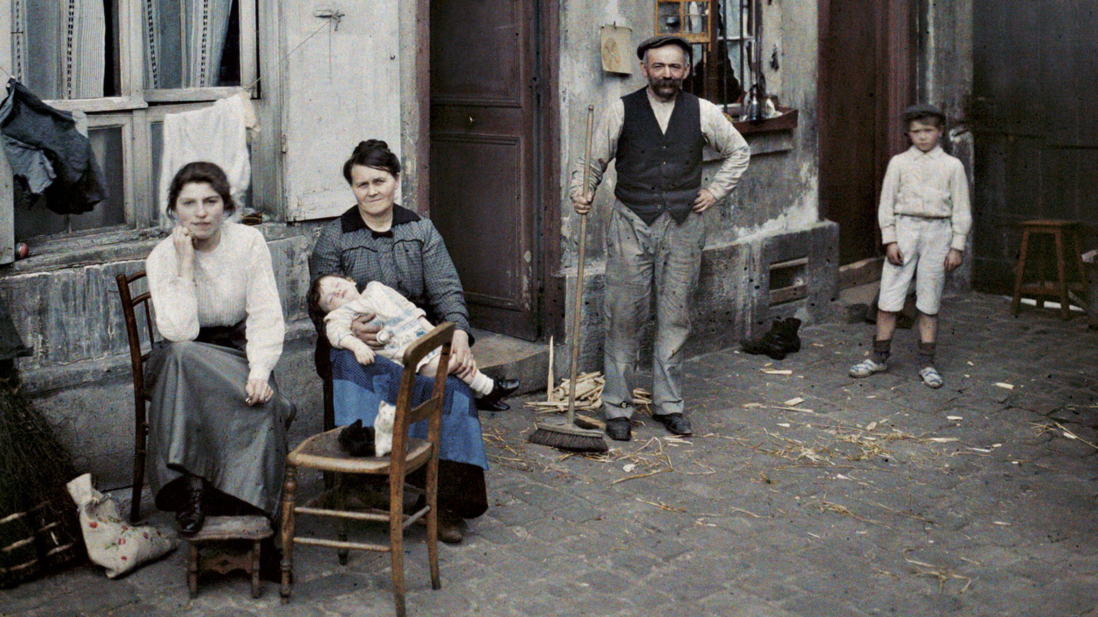 Early 20th-century colour photo of a family sitting and standing outside a rustic building on a cobblestone street.