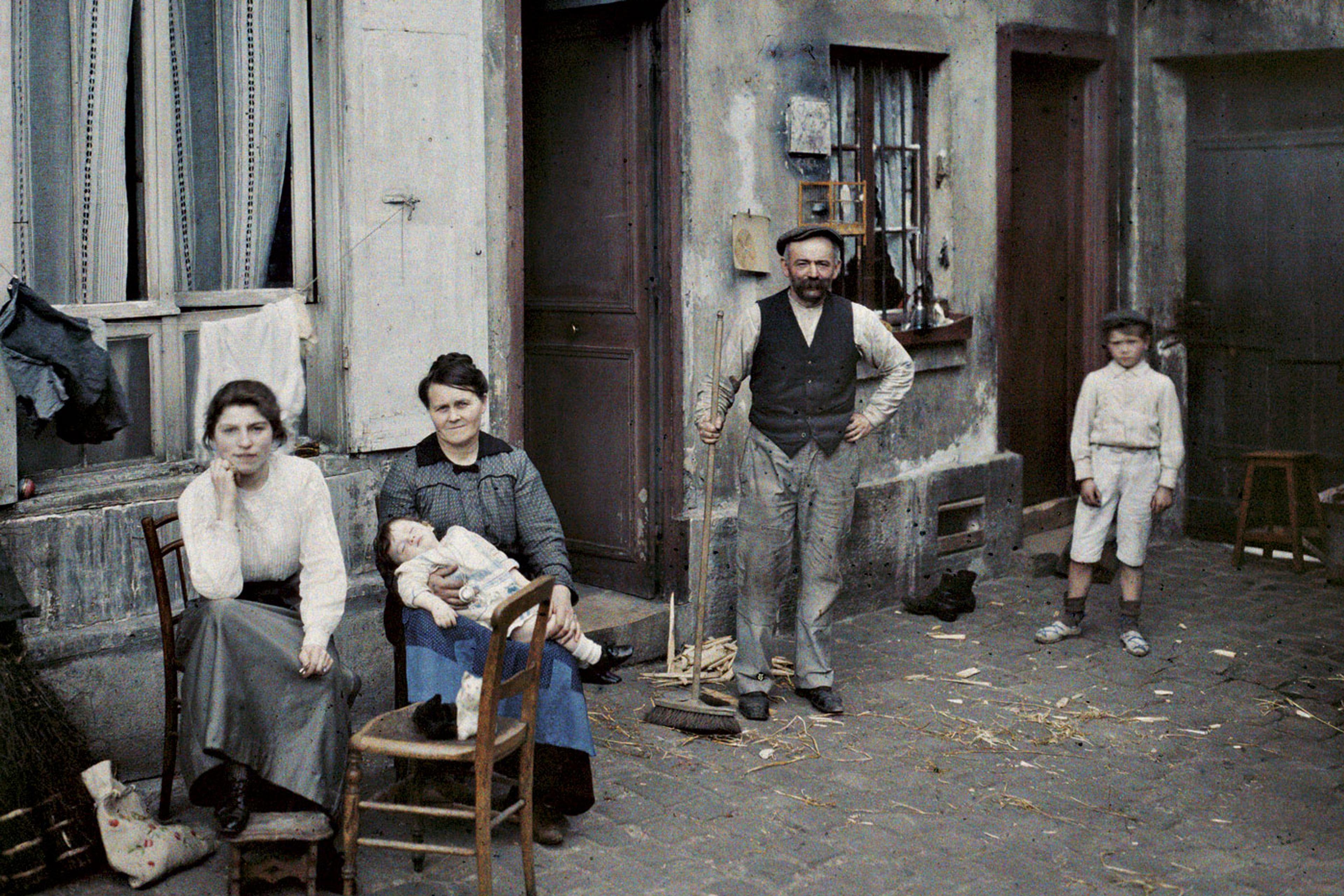 Early 20th-century colour photo of a family sitting and standing outside a rustic building on a cobblestone street.