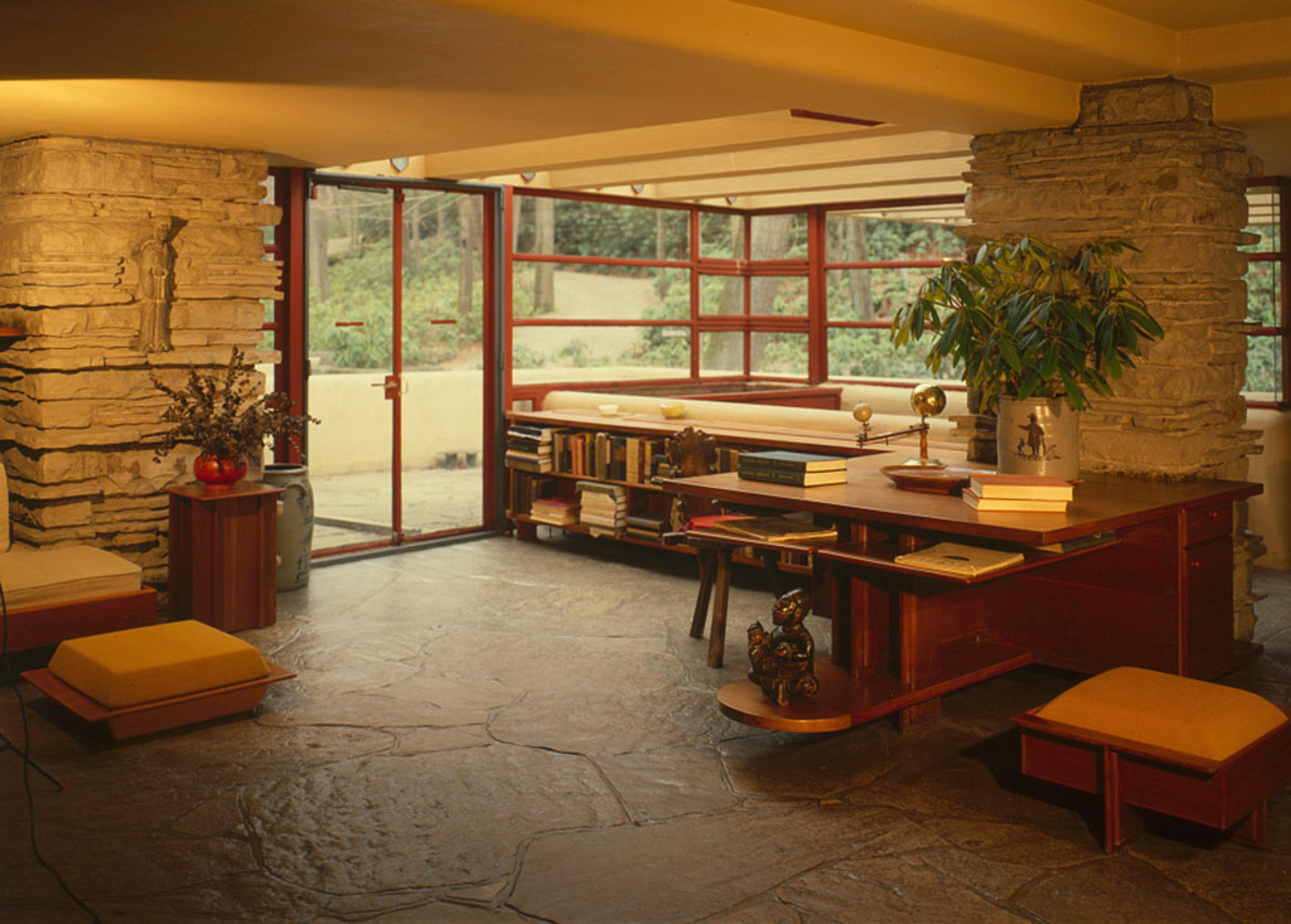 Interior photo of a room with stone walls and floor, wooden desk, books, plants and large windows with a garden view.