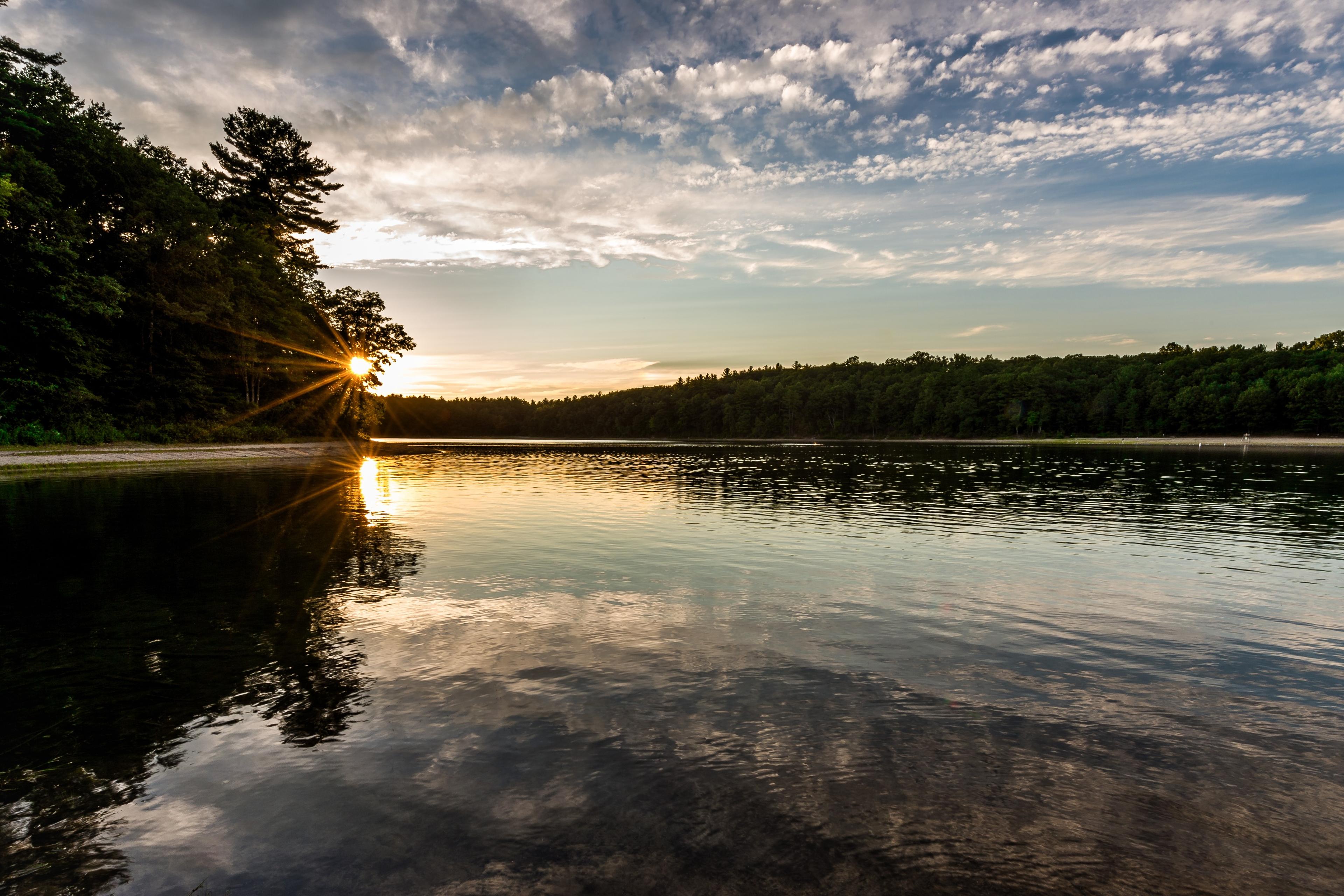 Photo of a serene lake at sunset with the sun peeking through trees, reflecting on the water and a cloudy sky above.