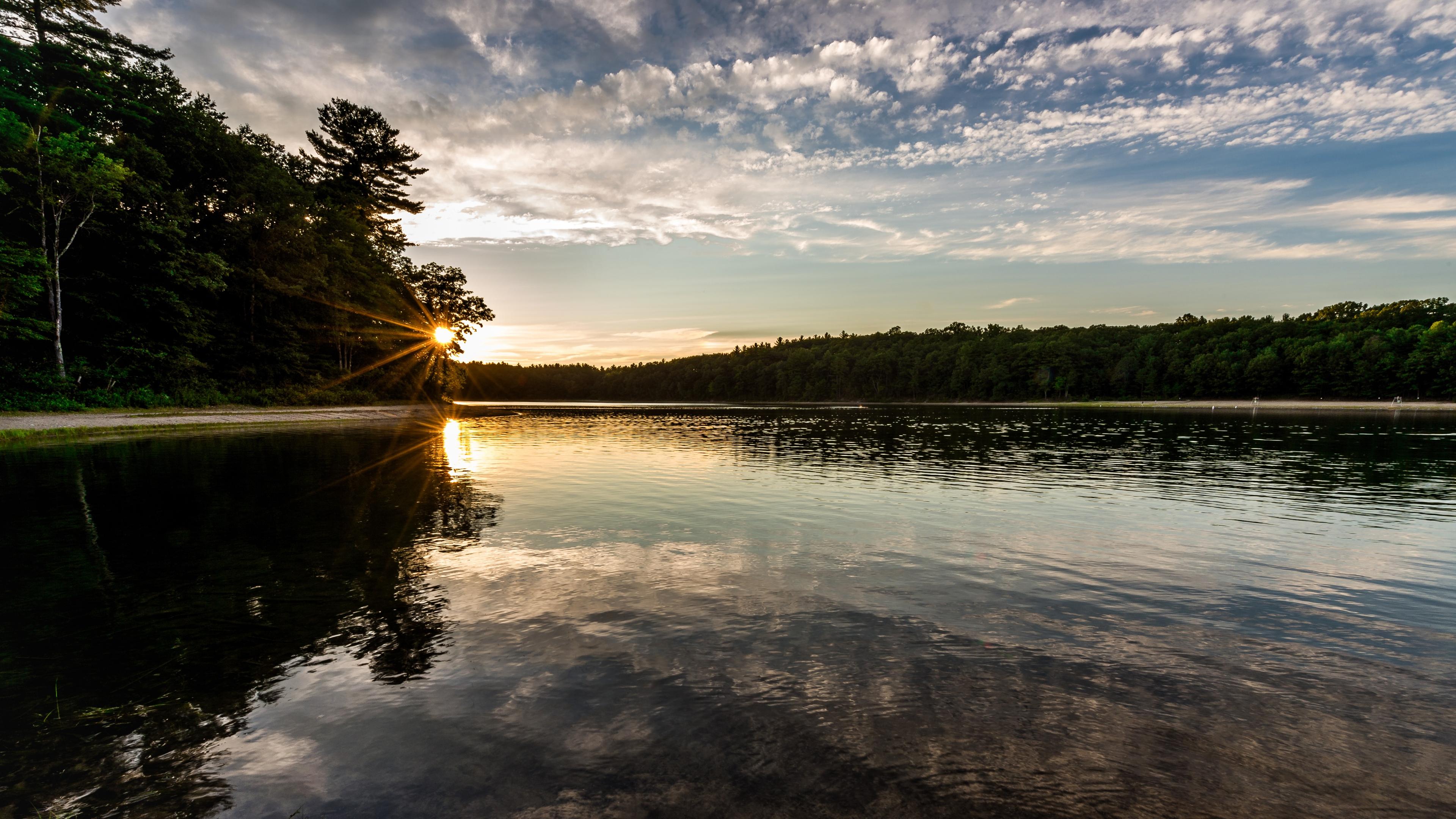 Photo of a serene lake at sunset with the sun peeking through trees, reflecting on the water and a cloudy sky above.