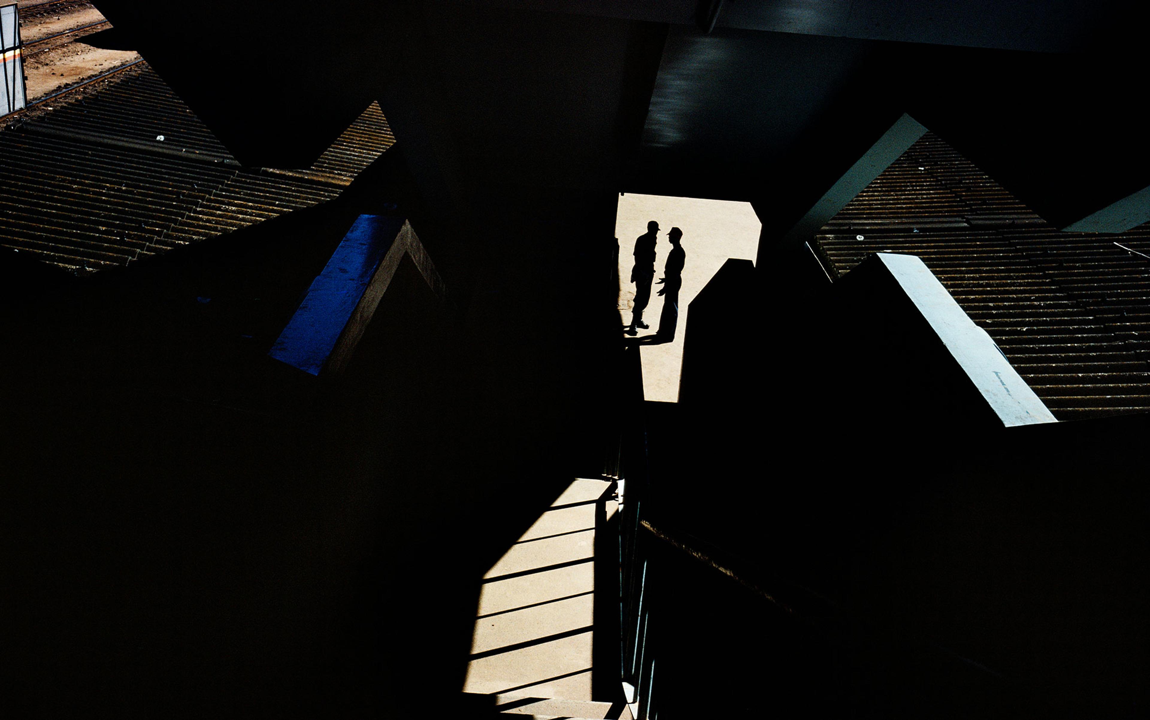 Photograph of two silhouetted figures on a sunlit path surrounded by dark geometric shadows and structures.