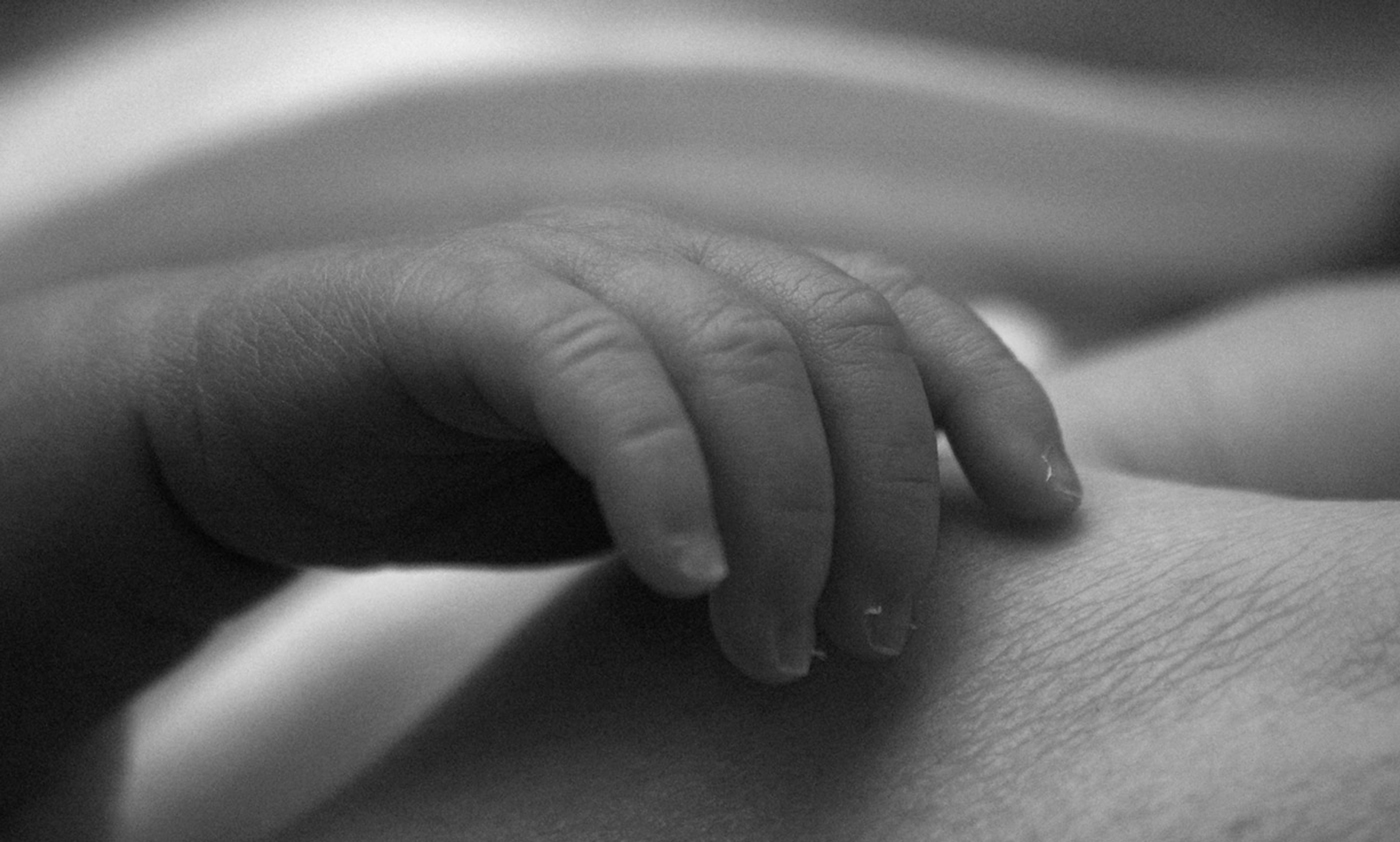 Black and white photo of a newborn baby’s hand resting on an adult’s hand.