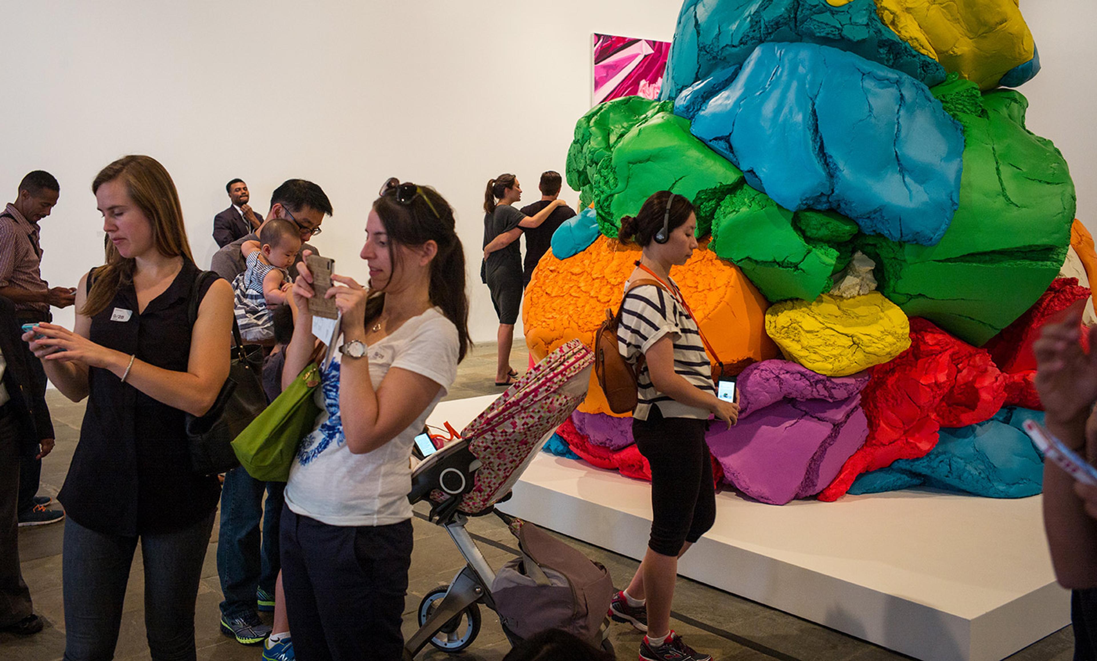 Museum visitors looking at phones near a large, colourful abstract sculpture. One visitor has a pram and another wears headphones.
