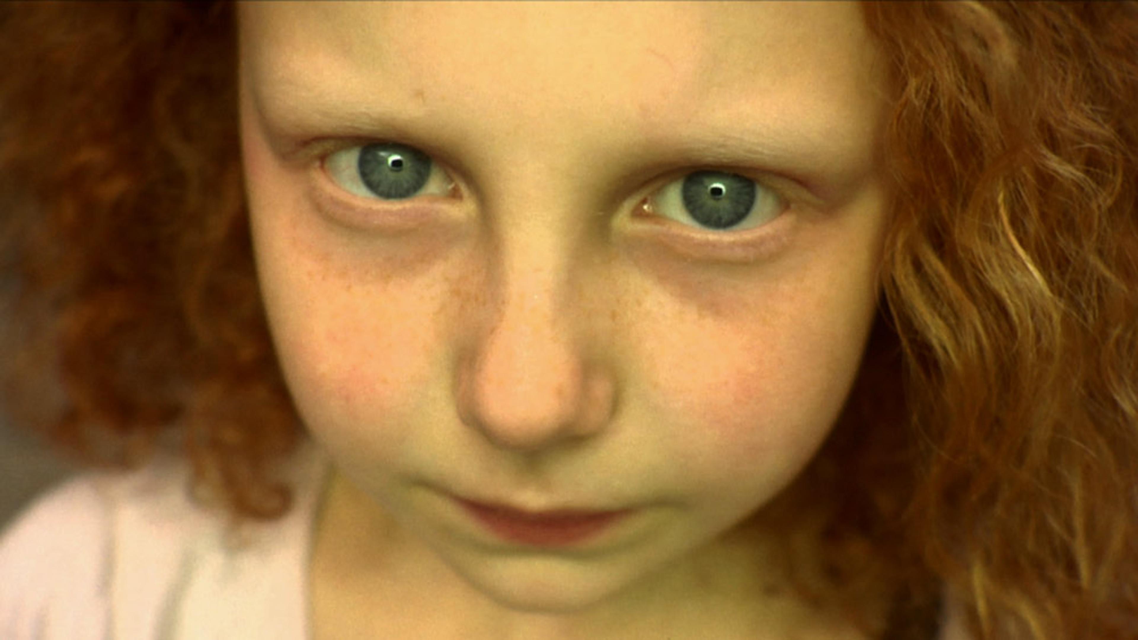 A close-up girl’s face with blue eyes and curly red hair, staring directly at the camera with a neutral expression.