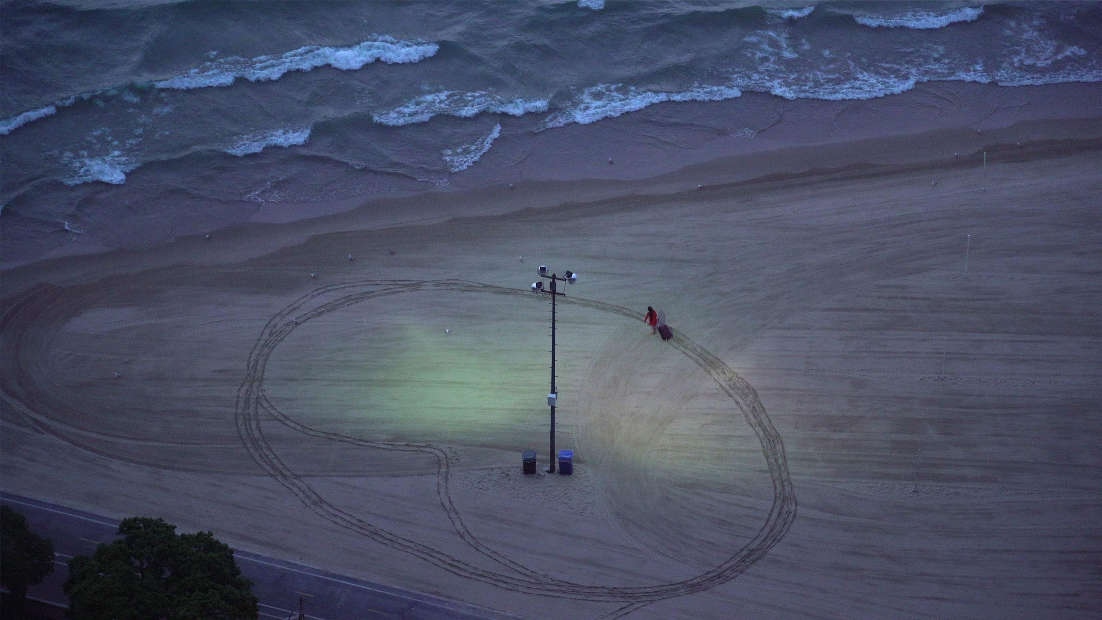 Aerial photo of a beach at dusk with a spotlight illuminating sand tracks seagulls waves and a person walking with luggage.