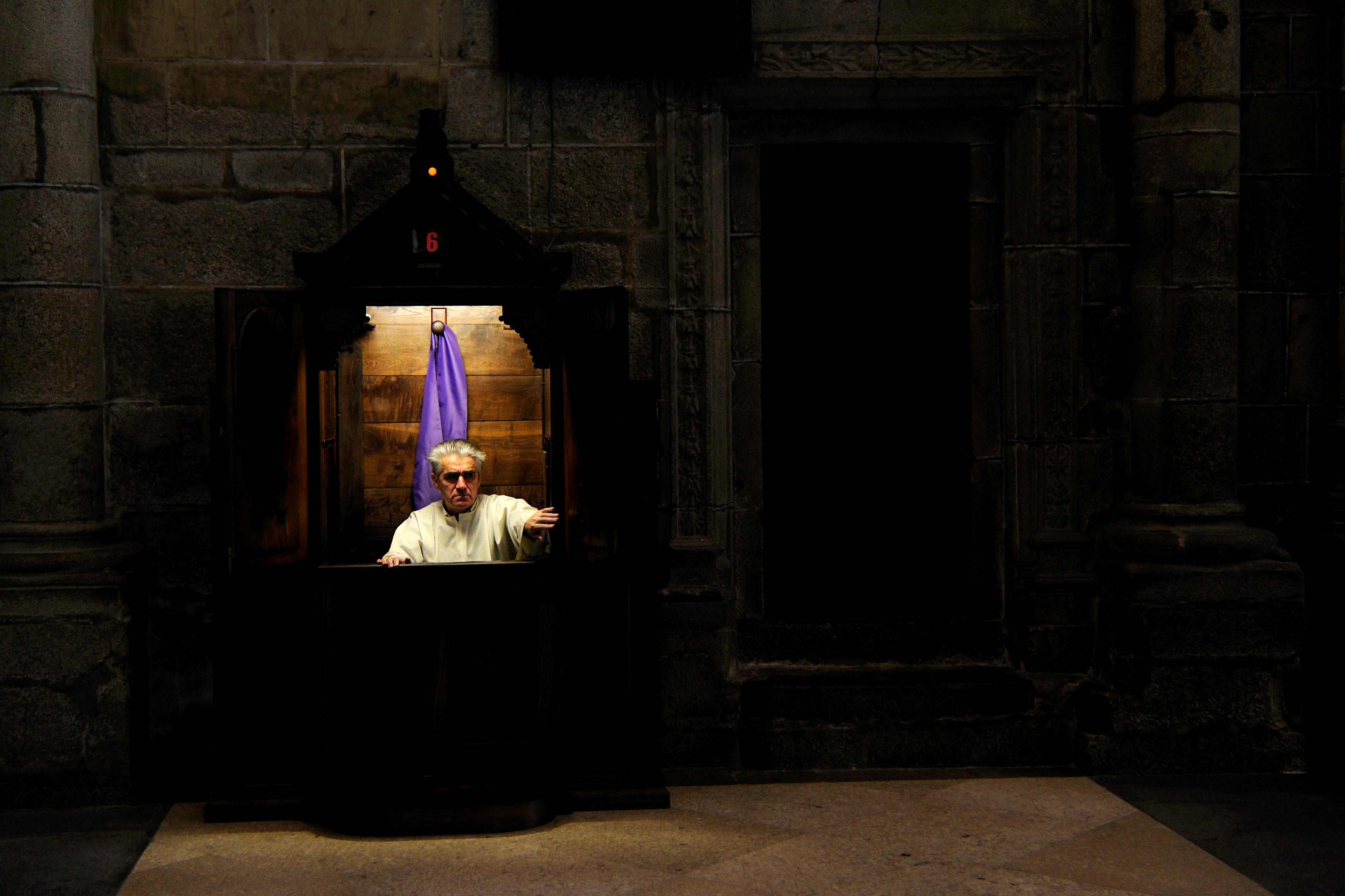 Photo of a man in a confessional booth with a purple curtain in a dimly lit stone-walled church setting.