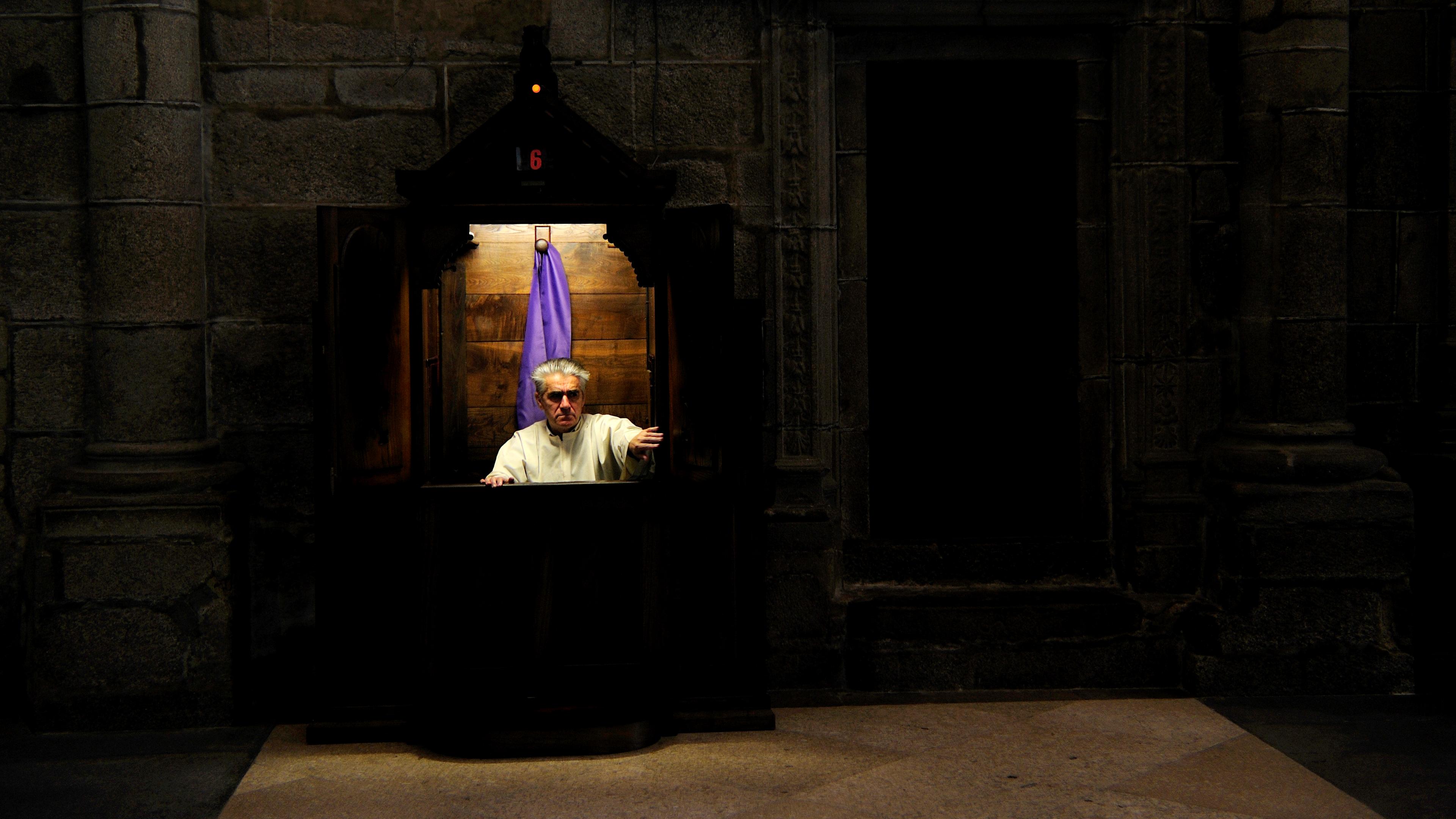 Photo of a man in a confessional booth with a purple curtain in a dimly lit stone-walled church setting.