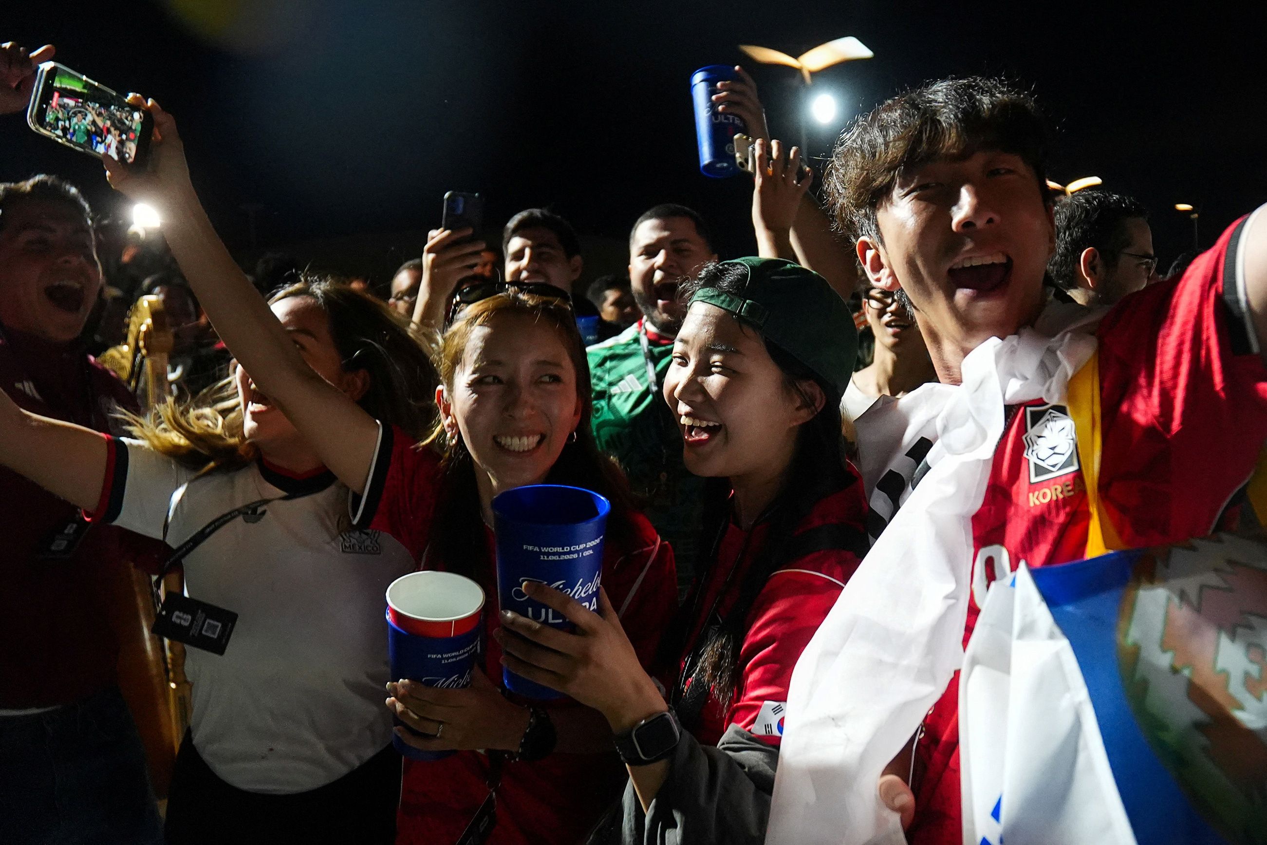 Excited football fans in team jerseys celebrating, some holding cups, others taking selfies at night.