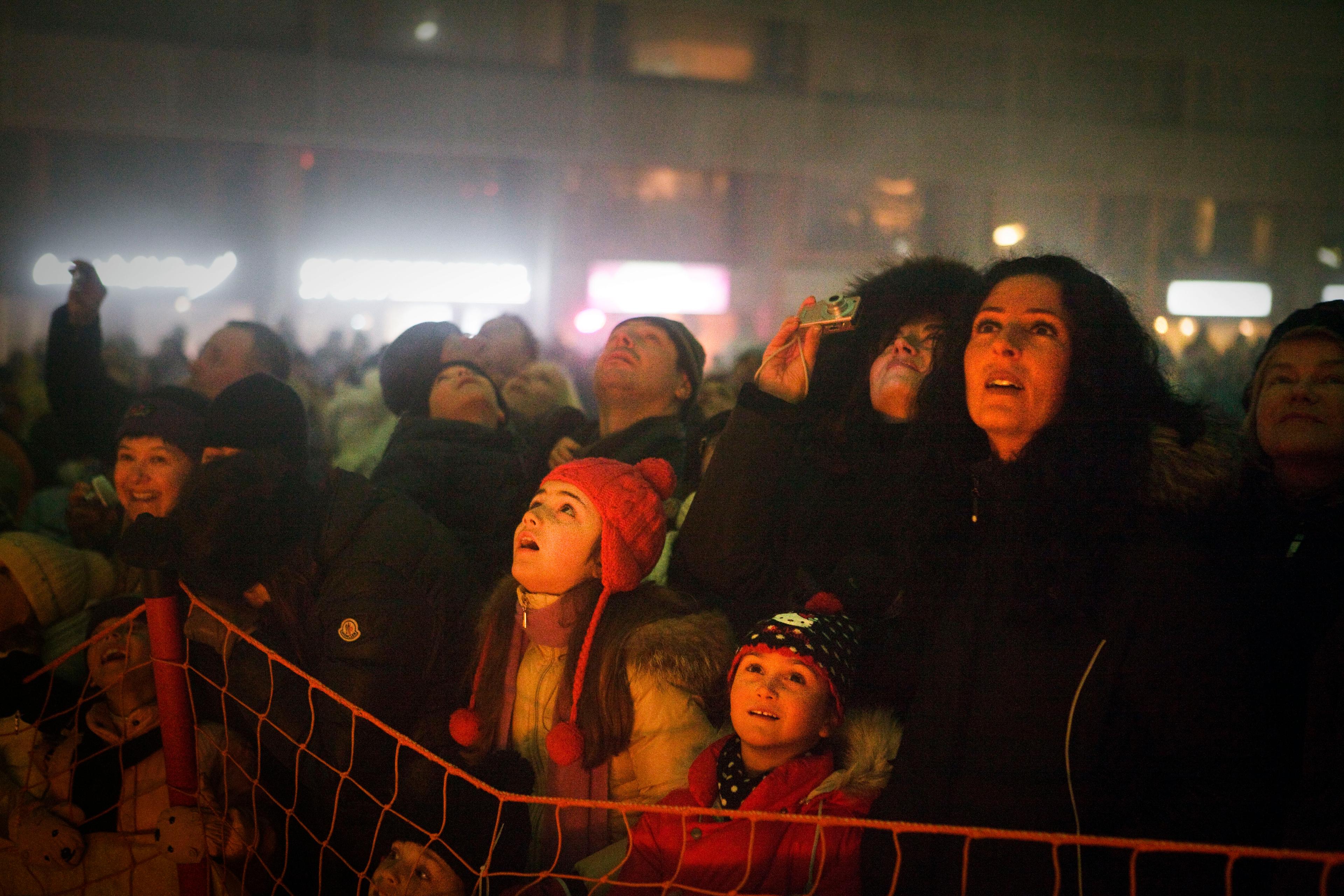 Photo of a crowd watching an event at night, faces lit, some taking photos, children in colourful hats by a barrier.