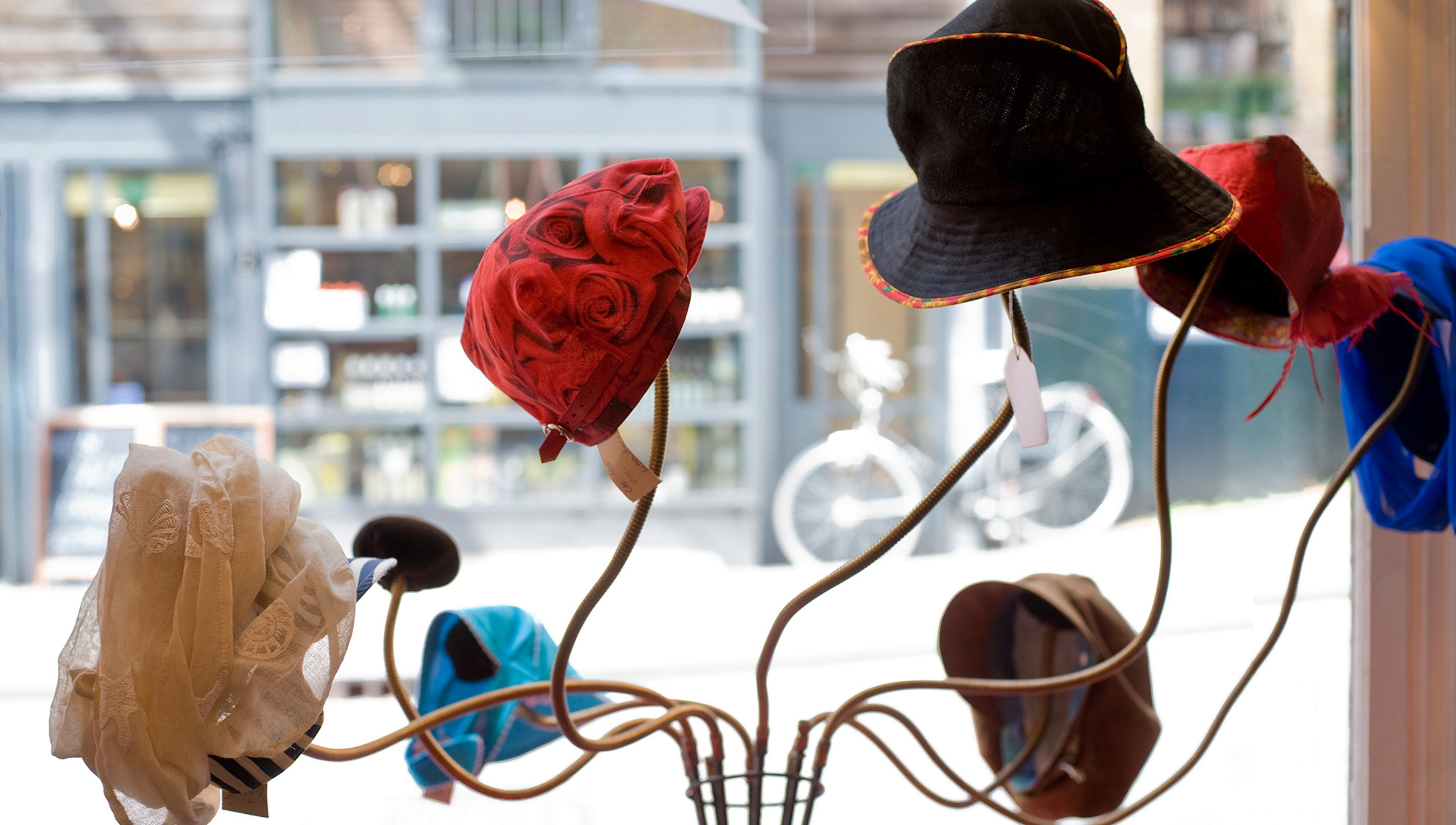 A display of various hats, including a red floral hat, a black hat with a coloured brim, a blue hat, and a beige lacy hat, shown on stand in a shop window. A blurred street with a bicycle is visible outside.
