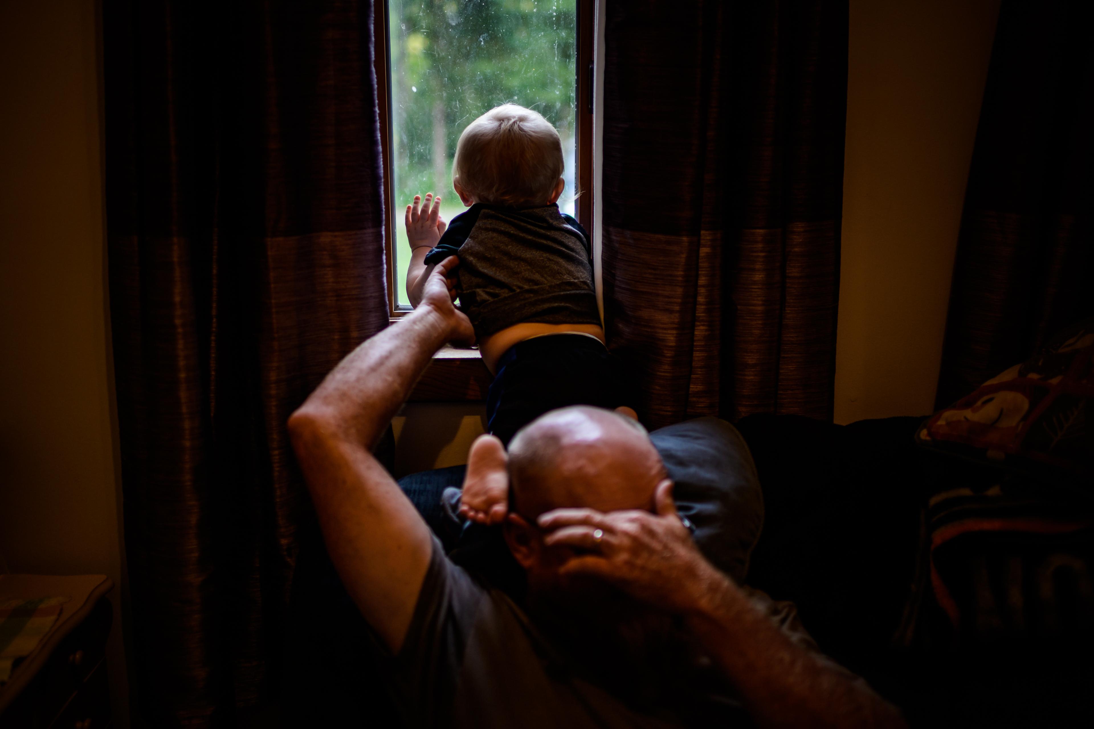 A child looking out a window, hand pressed against the glass, supported by an adult holding their back, in a warmly lit room with partially-closed curtains.