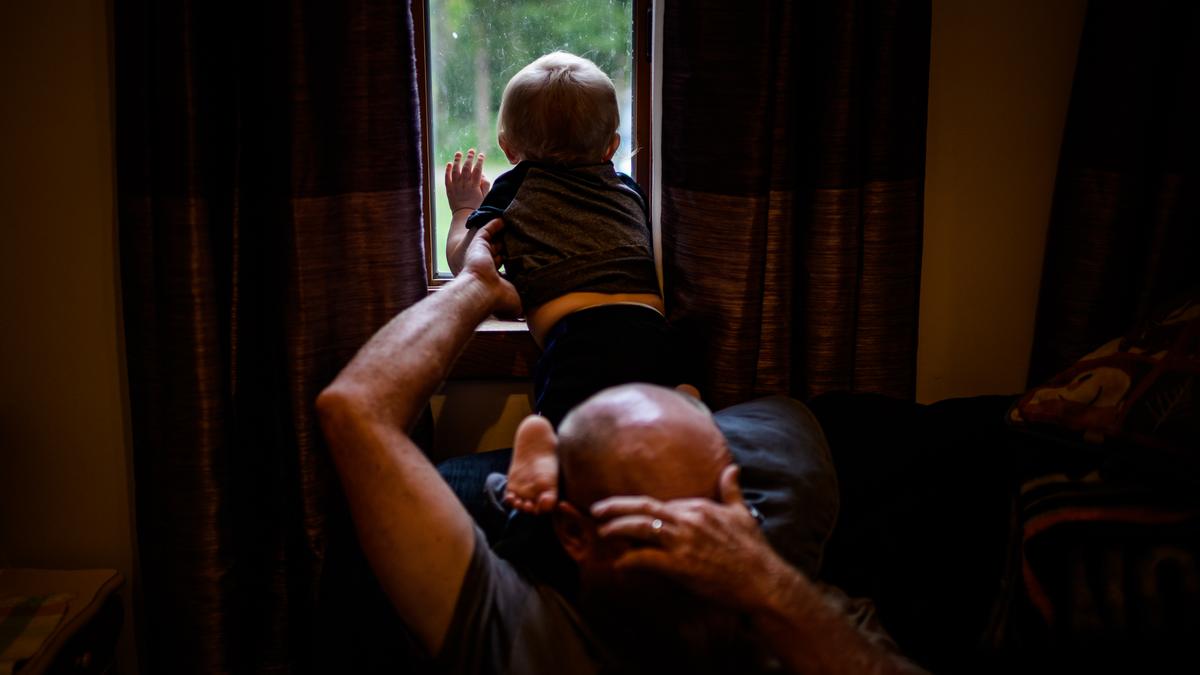 A child looking out a window, hand pressed against the glass, supported by an adult holding their back, in a warmly lit room with partially-closed curtains.