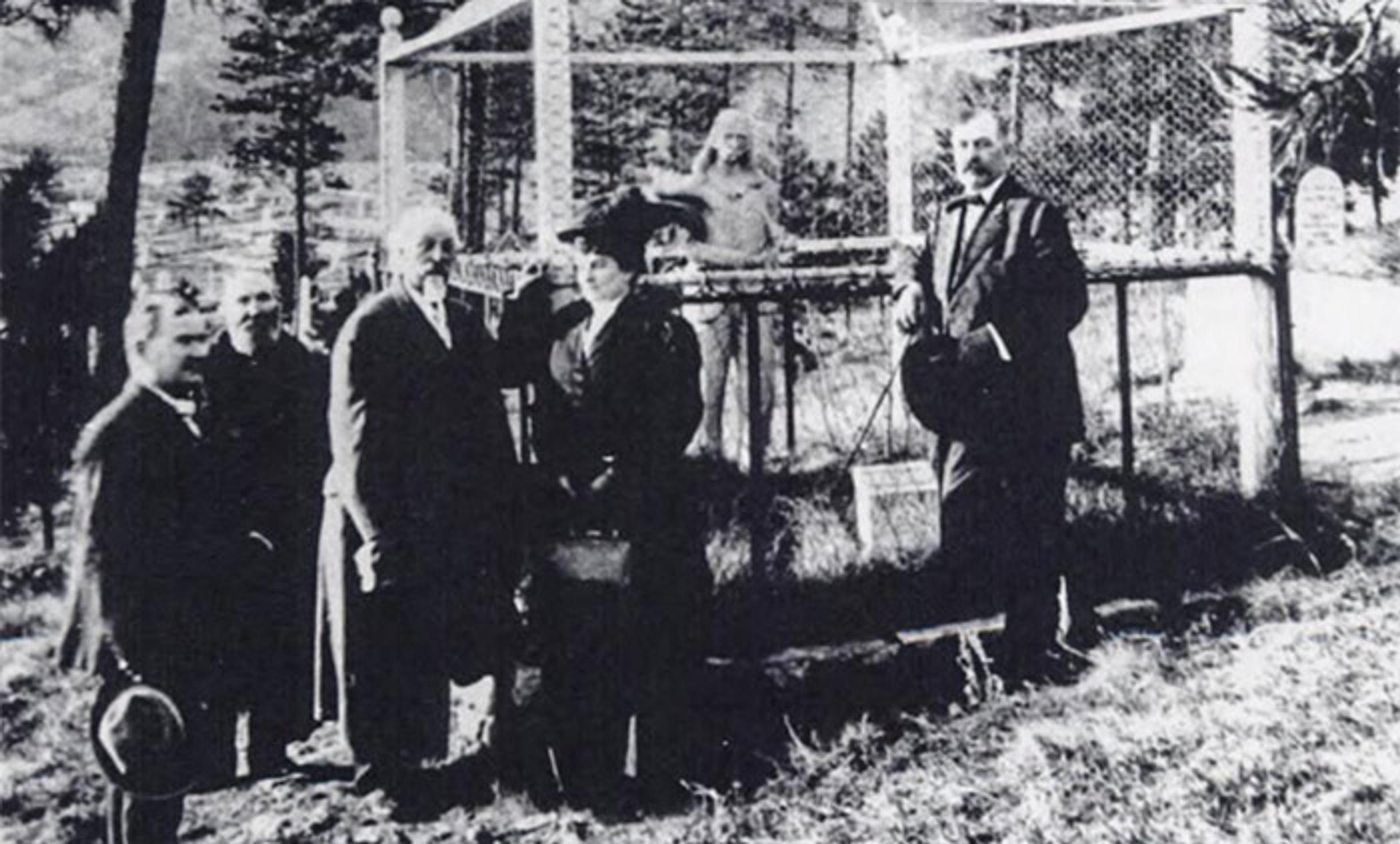 Vintage photo of people gathered outdoors around a cage with a man inside. Trees are visible in the background.
