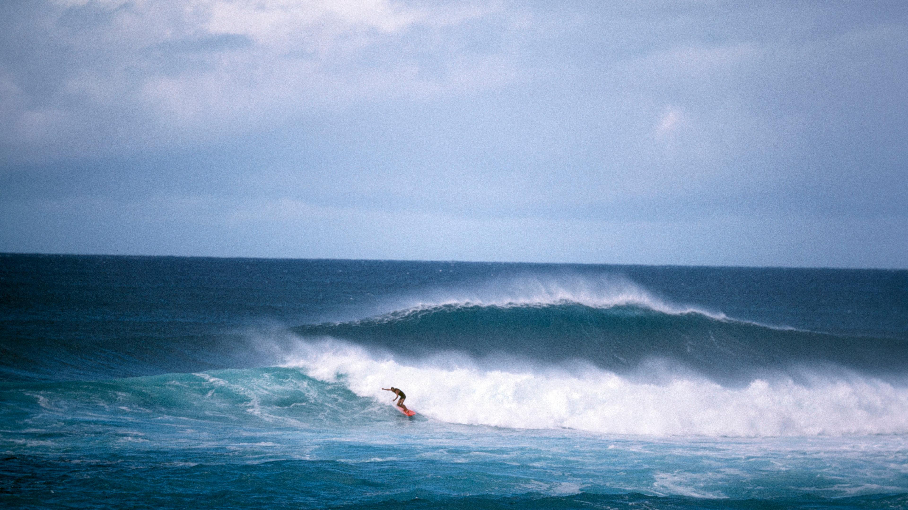 A surfer riding a wave under a cloudy sky, with another large wave shown behind.