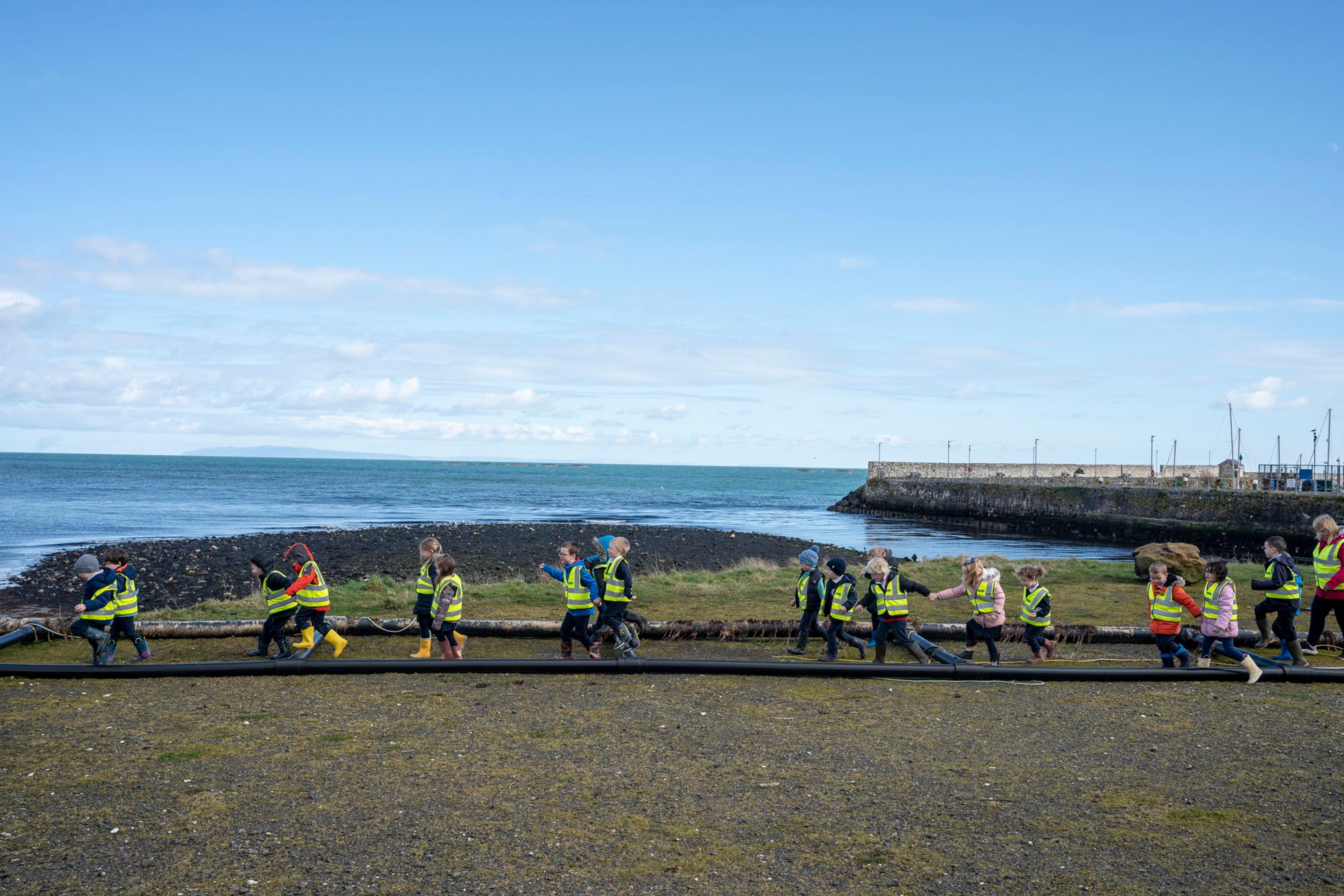 Photo of children in high-vis jackets walking along a grassy shore by the sea on a clear day.