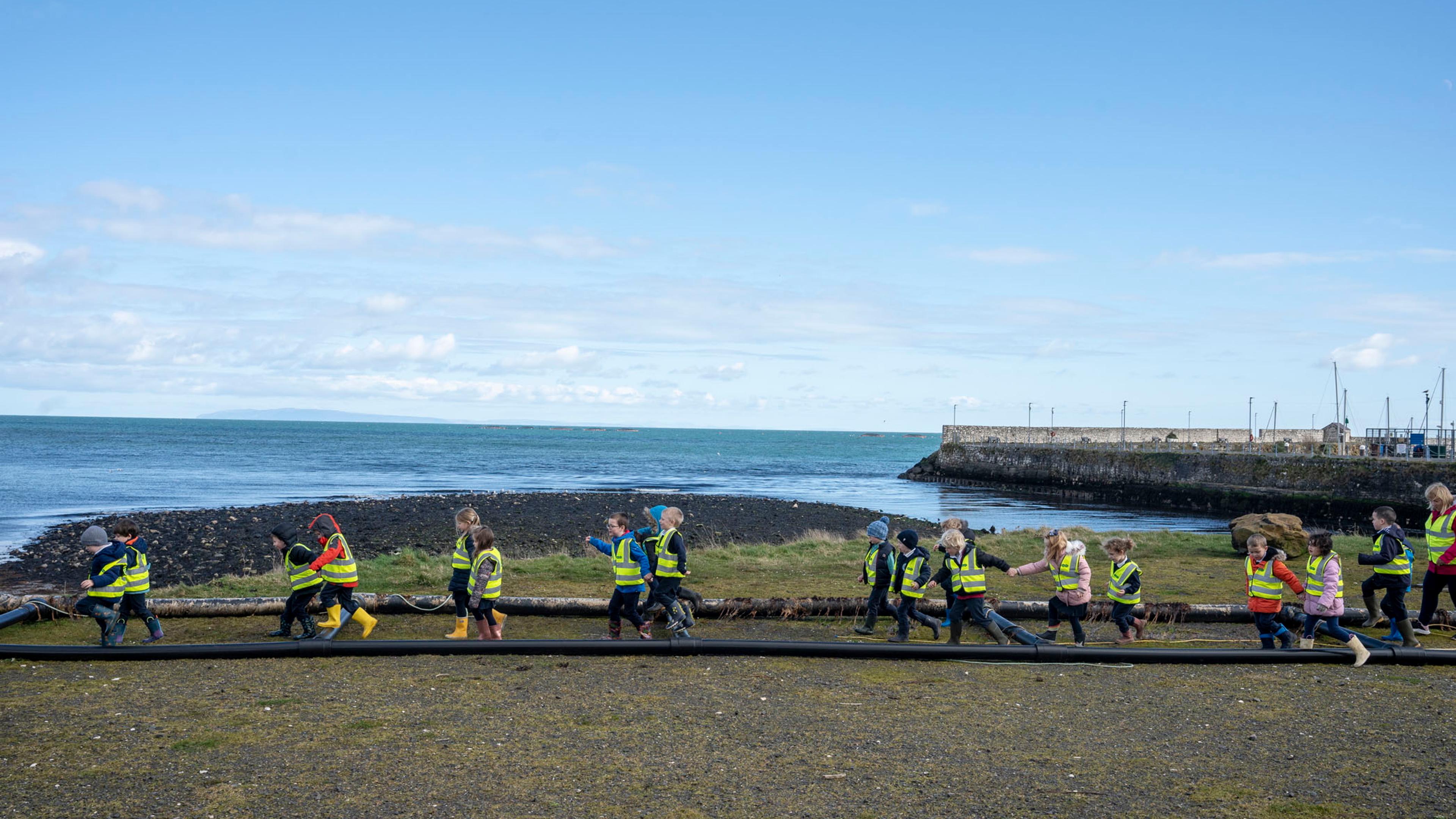 Photo of children in high-vis jackets walking along a grassy shore by the sea on a clear day.