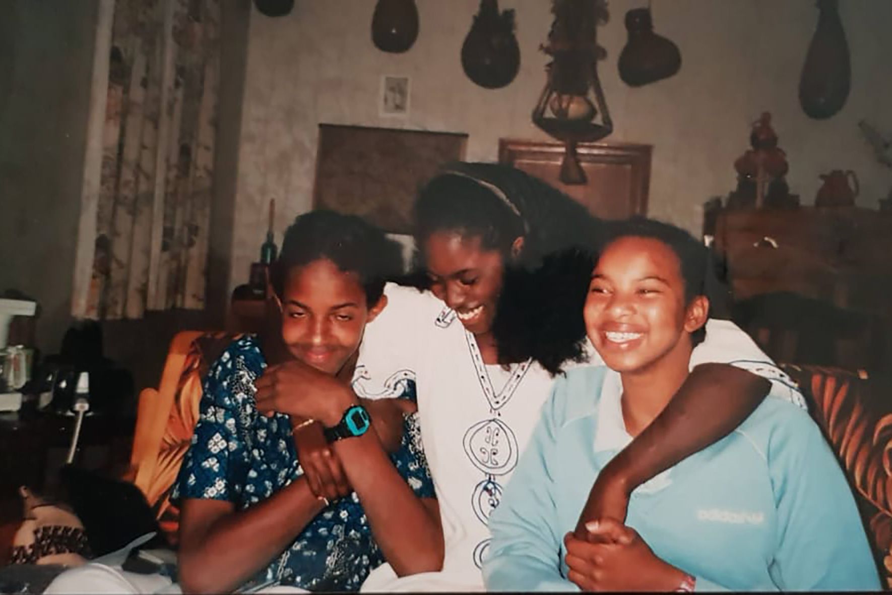 Photo of three people smiling indoors, seated with arms around each other, with decorative items in the background.