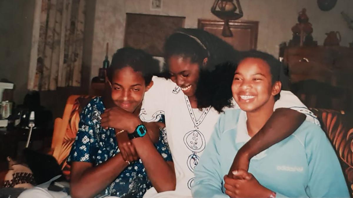 Photo of three people smiling indoors, seated with arms around each other, with decorative items in the background.