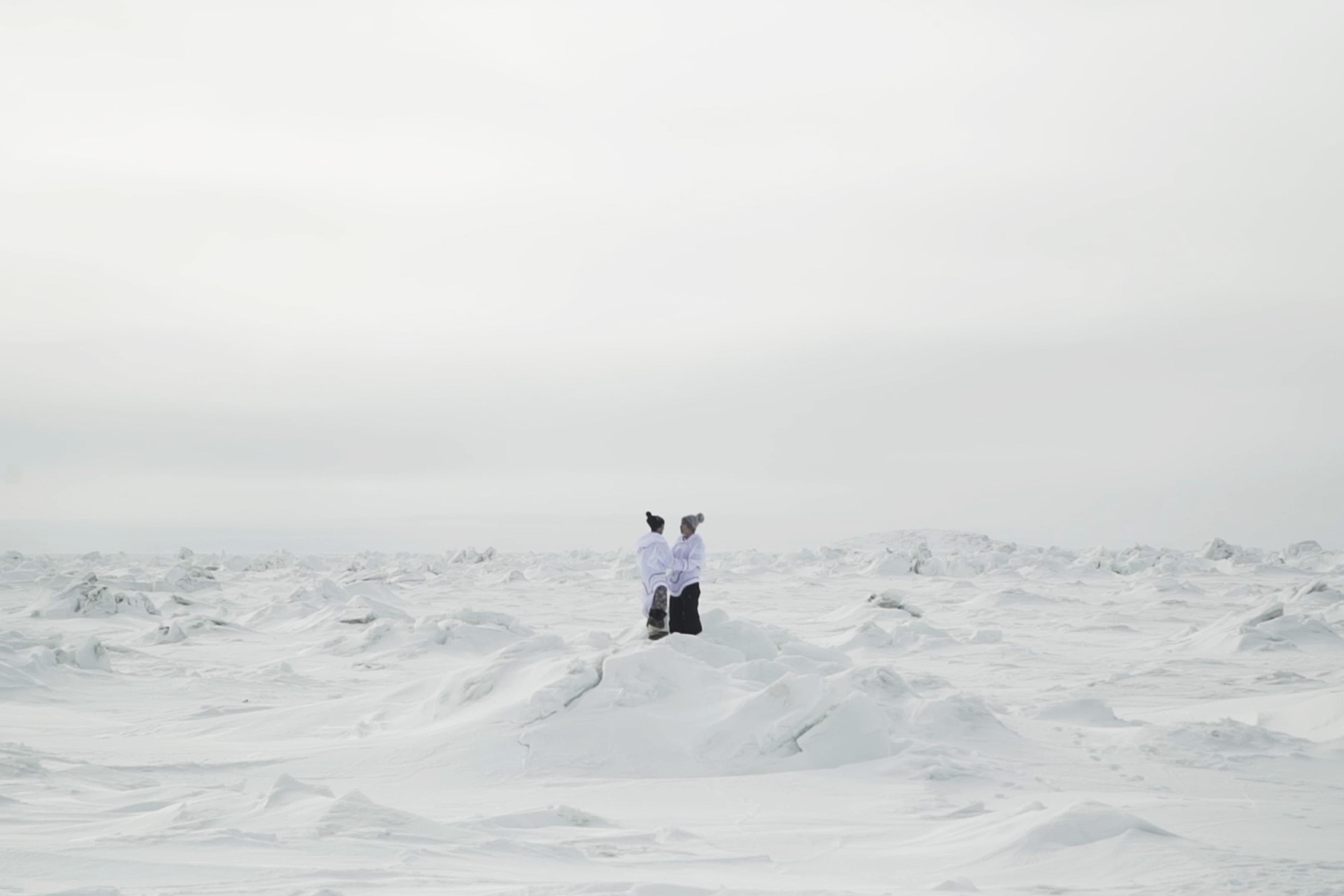 Two people wearing white jackets stand on a snowy landscape under an overcast sky, surrounded by snow-covered mounds.