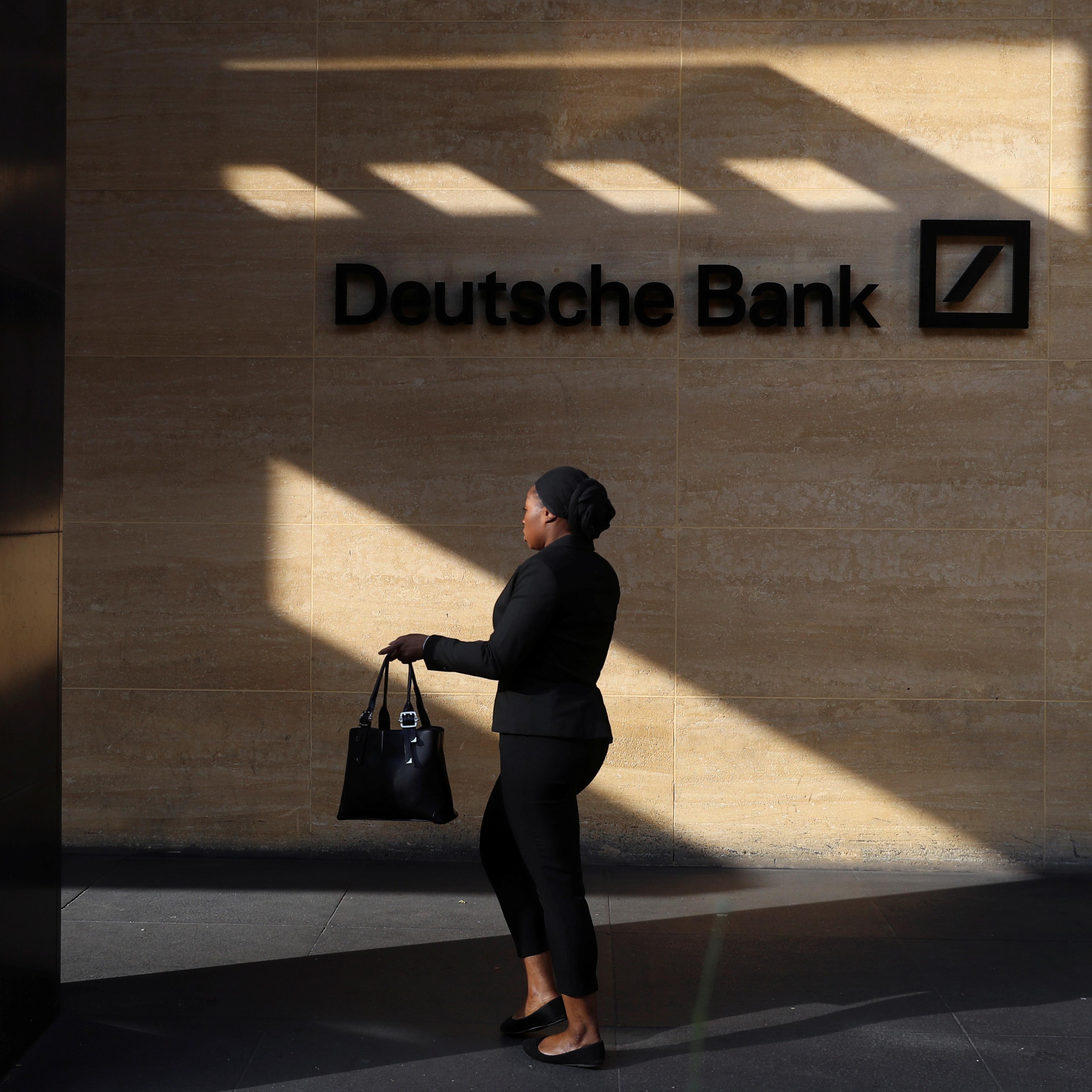 Photo of a person walking by a Deutsche Bank sign on a sunlit wall, casting a shadow.