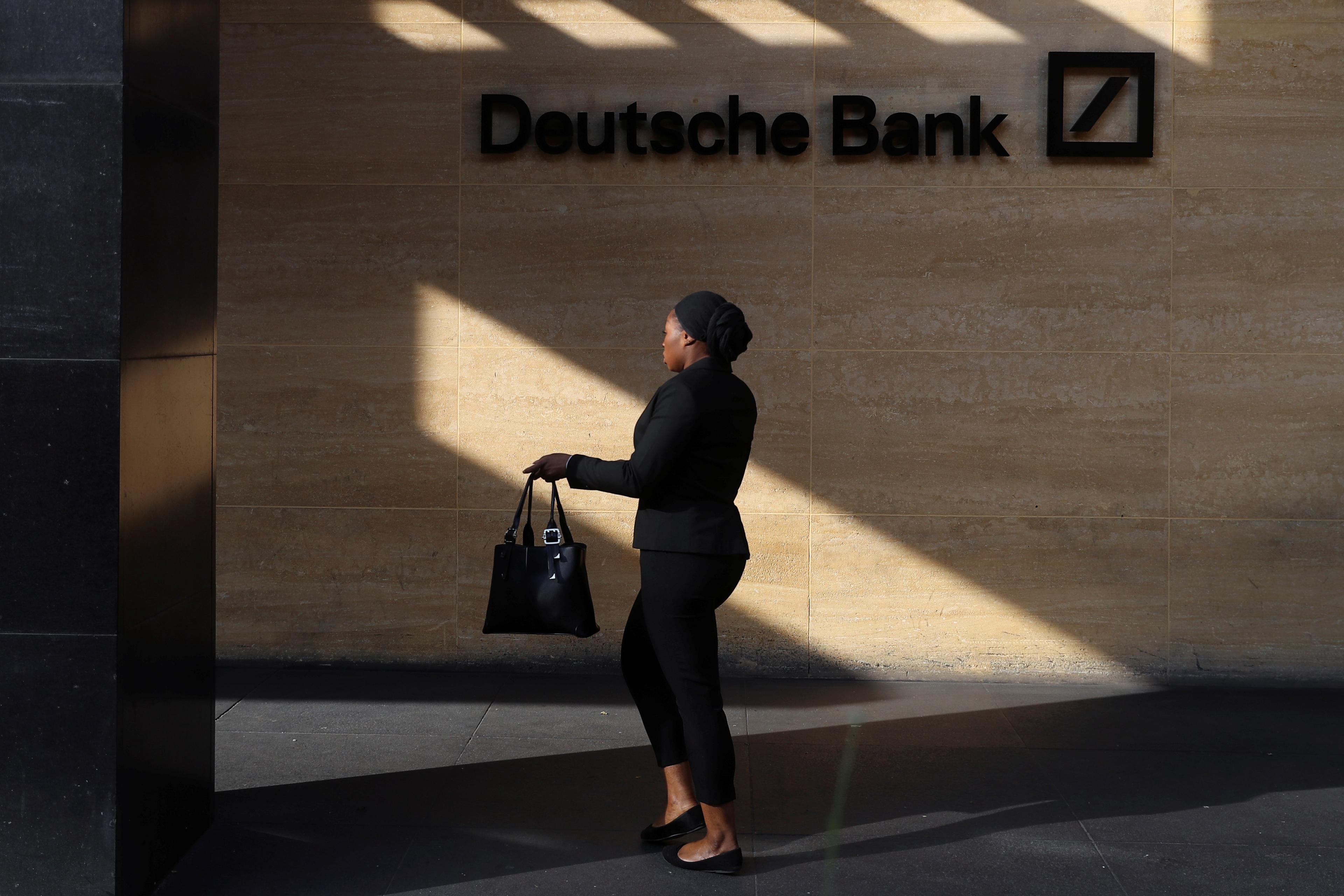 Photo of a person walking by a Deutsche Bank sign on a sunlit wall, casting a shadow.