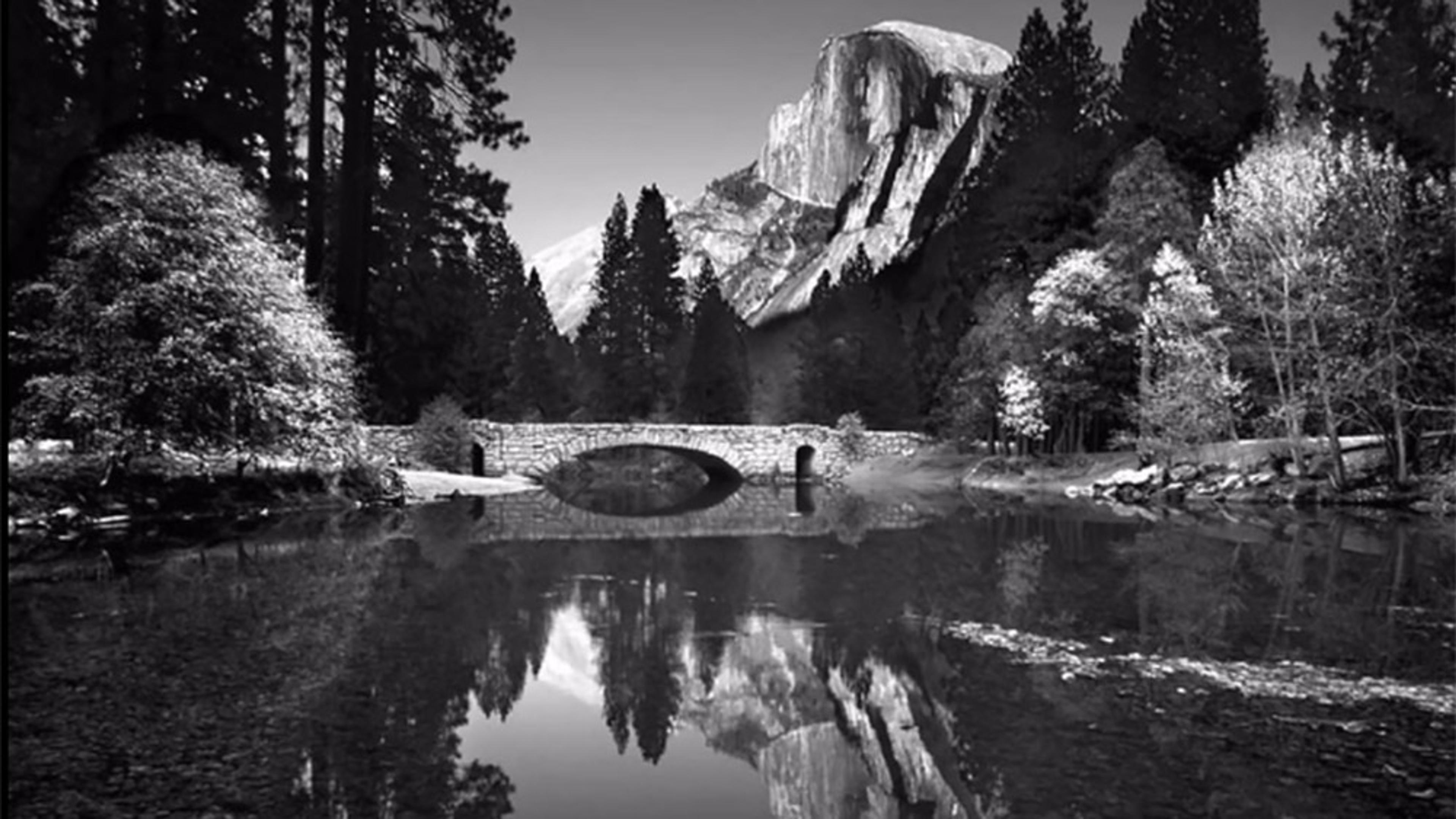 A black and white photo of a stone bridge over calm water with trees and a mountain reflected in the surface.