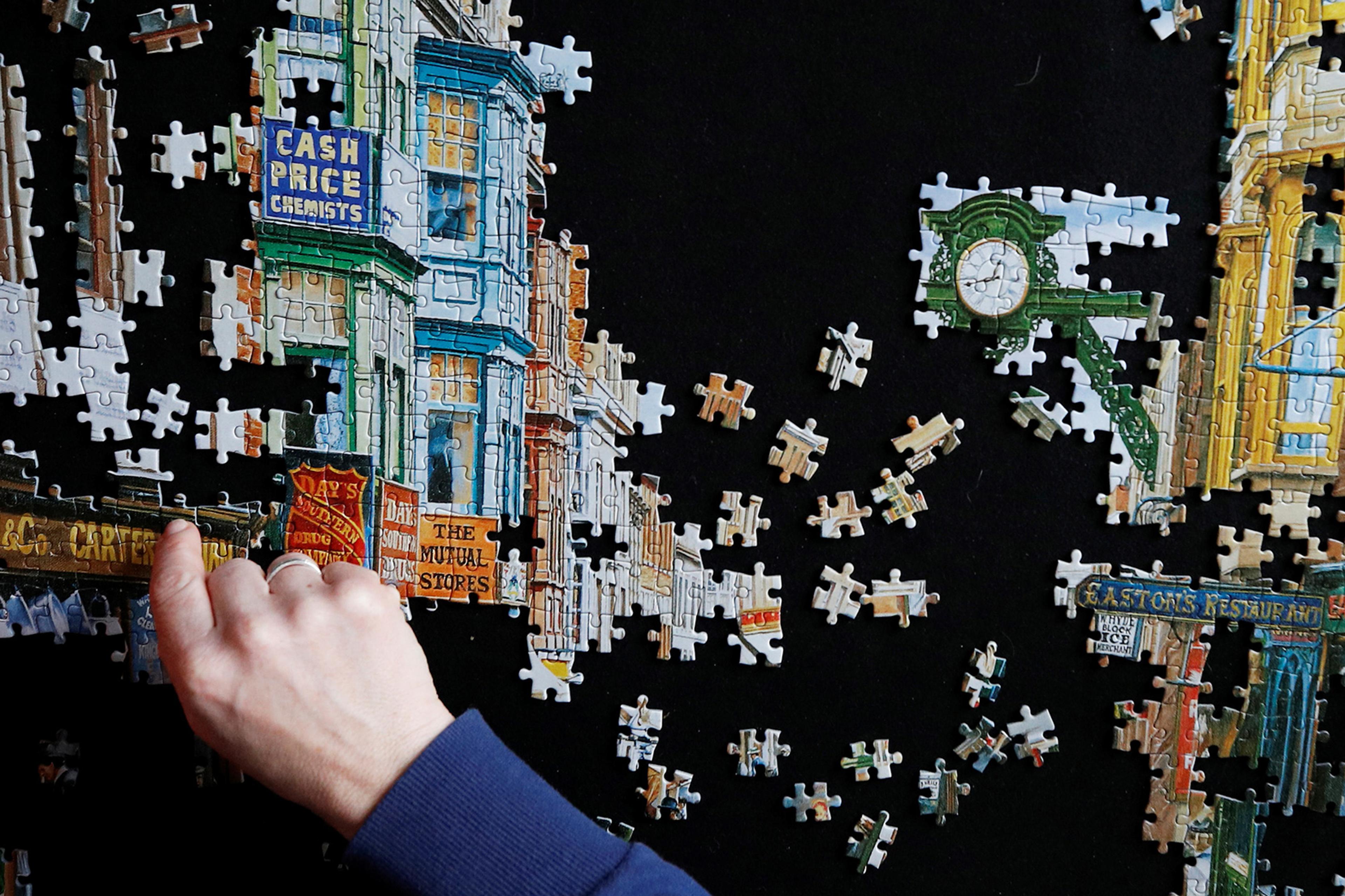 A close-up of a hand completing a jigsaw puzzle showing a street scene with shops and a clock against a black background.