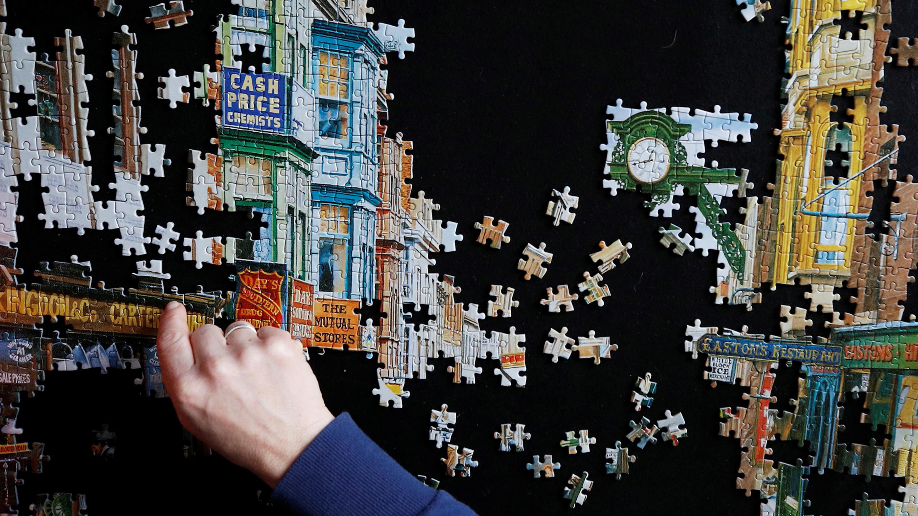 A close-up of a hand completing a jigsaw puzzle showing a street scene with shops and a clock against a black background.