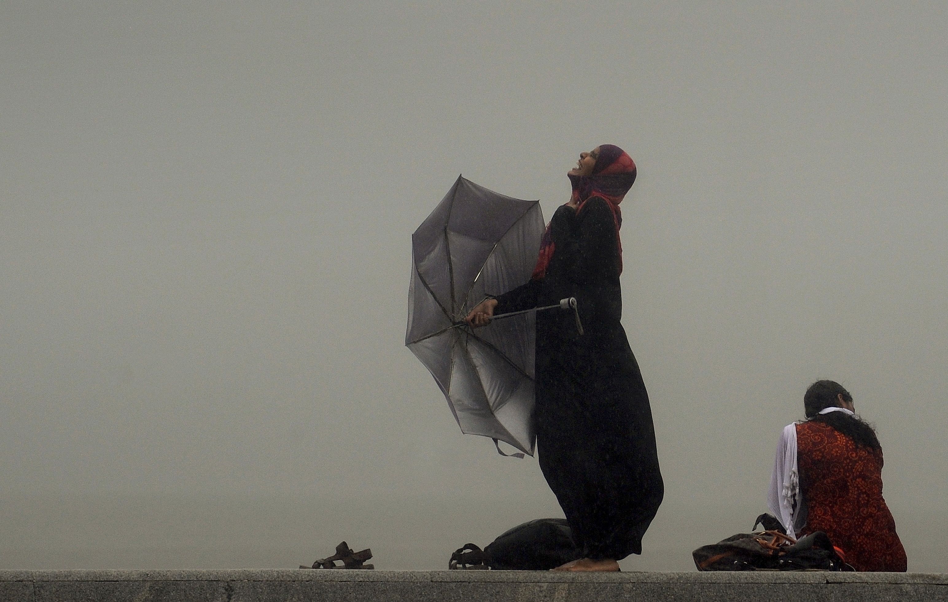 Photo of a woman in the rain holding a broken umbrella, smiling upwards; another sits nearby with a foggy background.